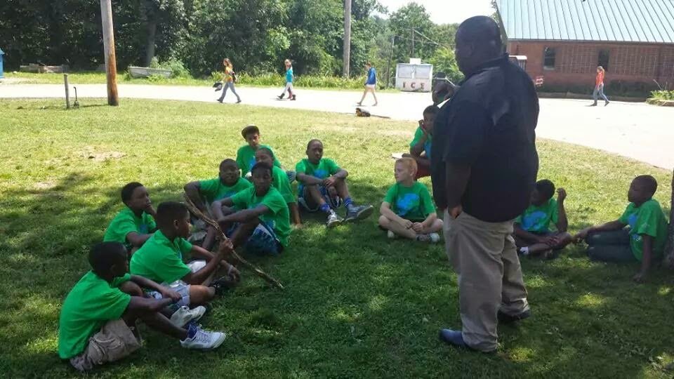 Group of children wearing green shirts sitting on grass in a circle outdoors, with adults standing nearby, under trees on a sunny day.