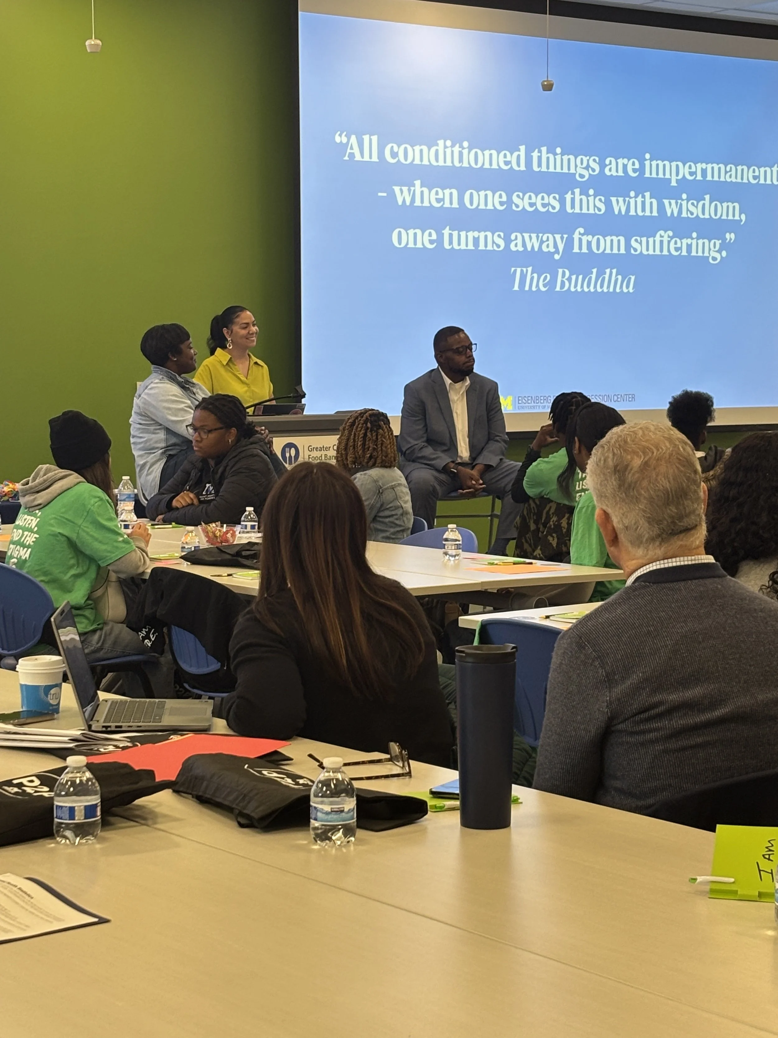 A group of diverse people in a meeting or conference room, with some looking at a projected quote on the wall that reads, 'All conditioned things are impermanent - when one sees this with wisdom, one turns away from suffering.' The room has green and blue walls, tables with water bottles, laptops, and papers, and several individuals are listening or speaking.