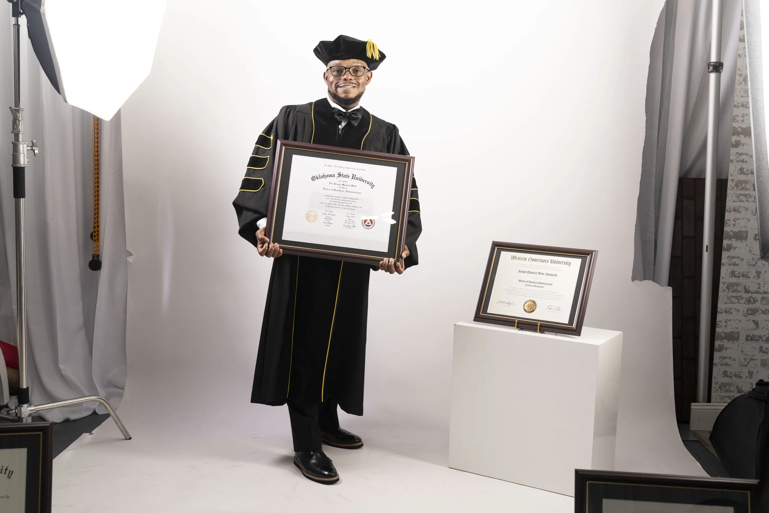 Graduate in black cap and gown holding diploma, standing in photo studio with framed diploma on pedestal to his right.