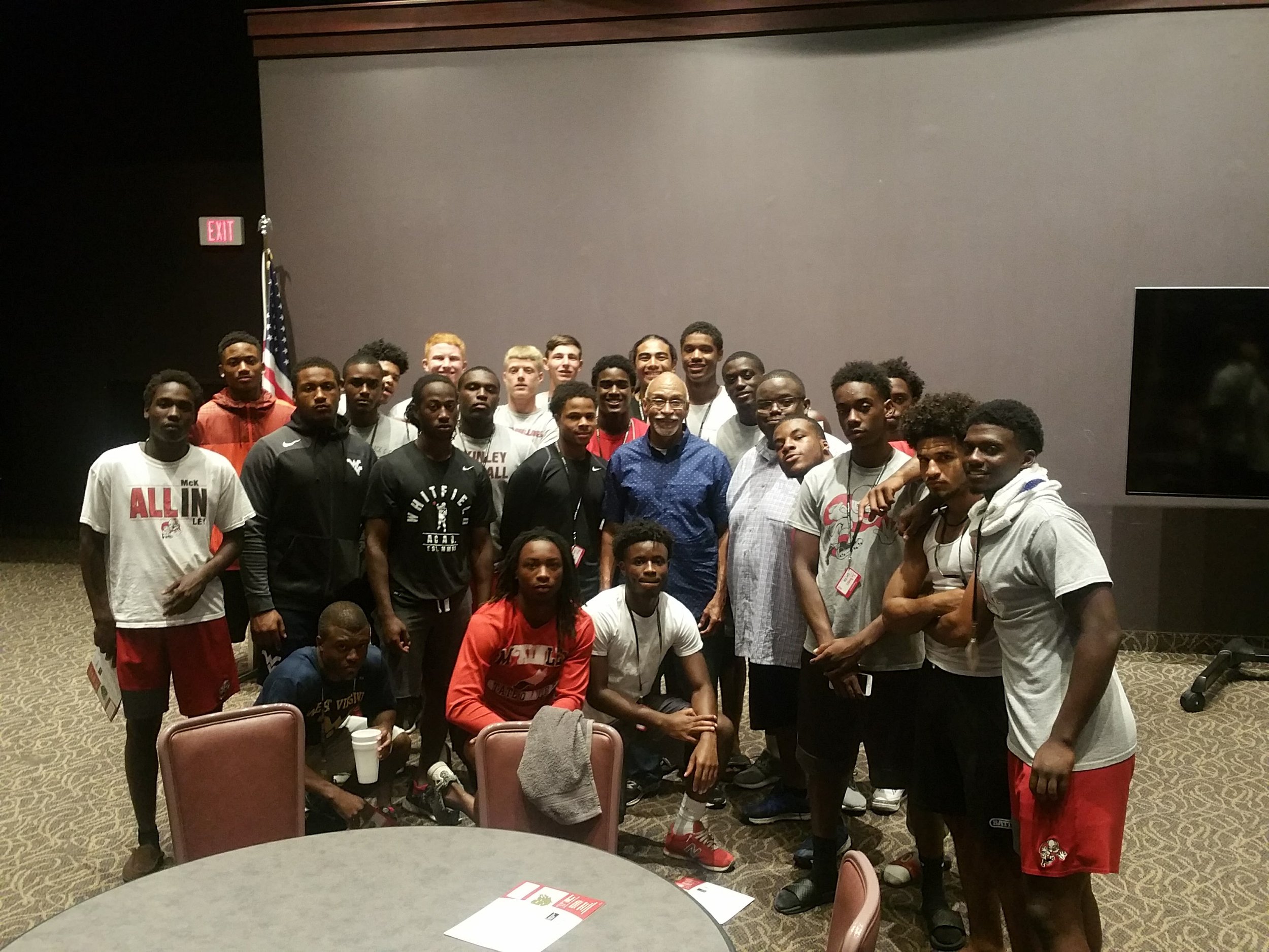 A group of young male athletes and a man in a blue shirt posing for a photo indoors, with some wearing athletic gear and T-shirts, and a large screen and American flag in the background.