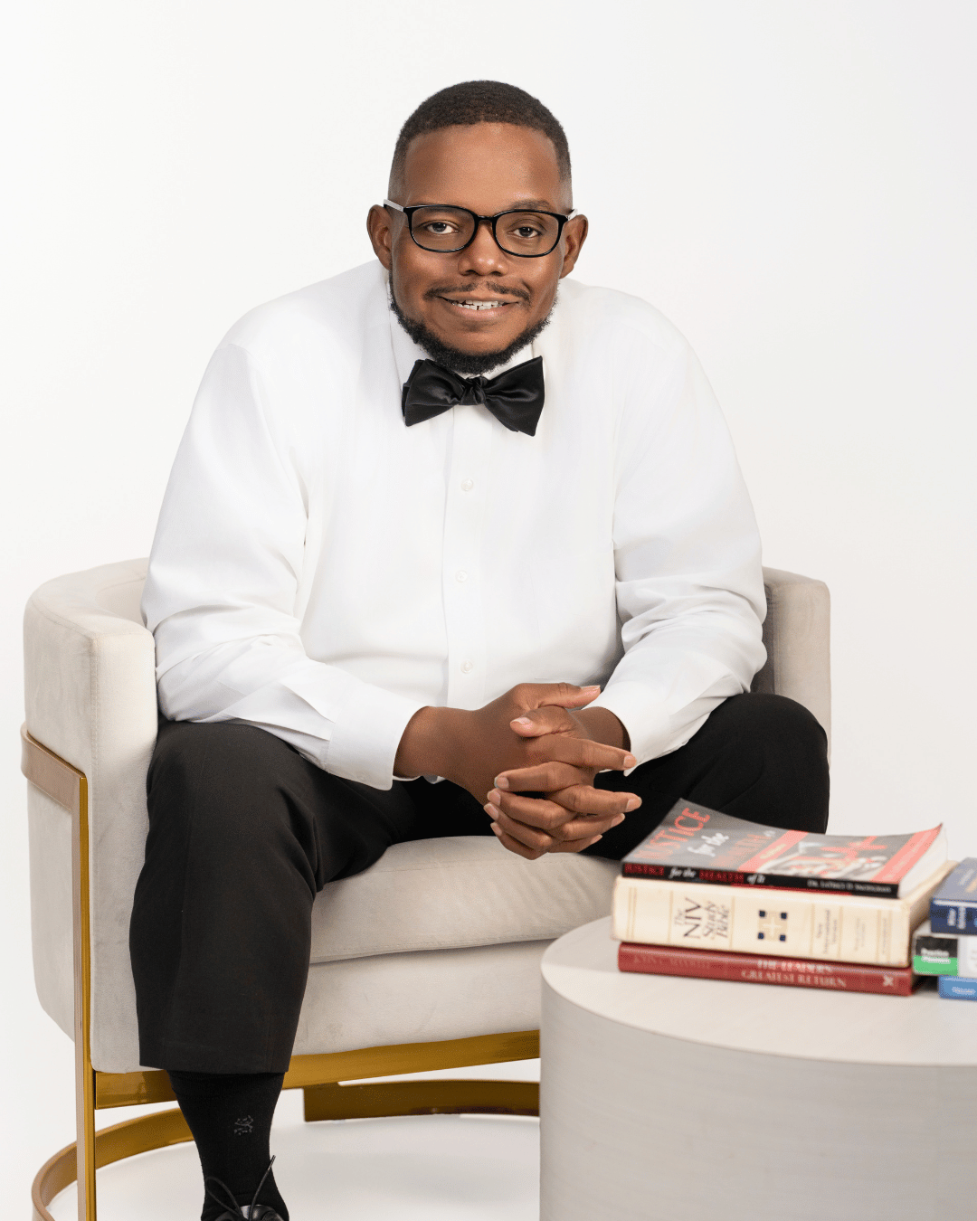 A man dressed in a white shirt and black bow tie sitting on a white armchair with a stack of books on a round table in front of him, against a plain white background.