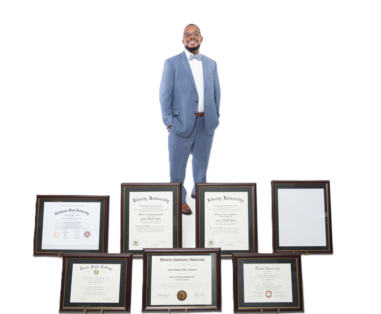 A man in a blue suit, glasses, and a bow tie standing with his hands in his pockets, smiling, behind a display of various framed diplomas and certificates.