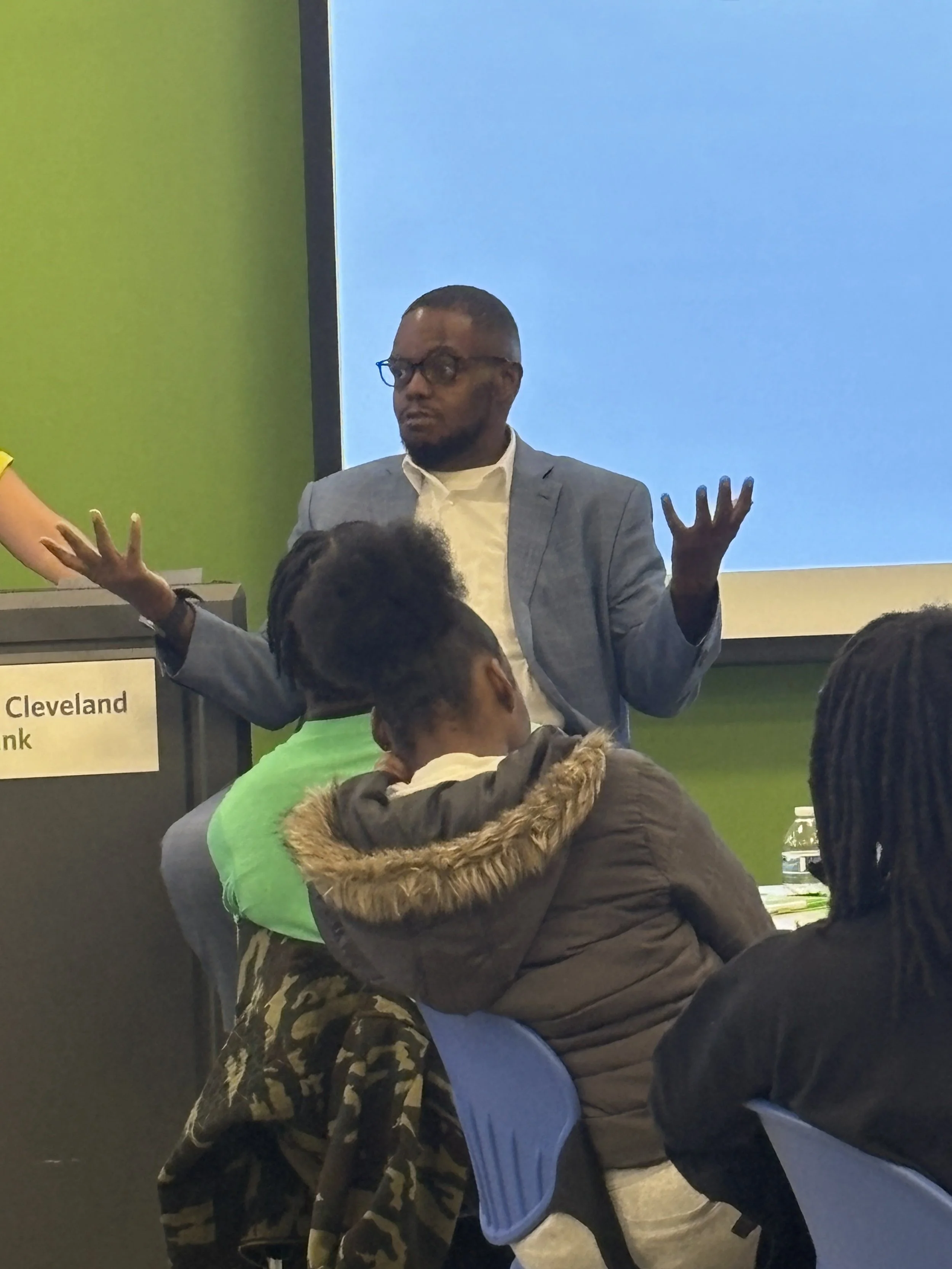 A man in a grey blazer and glasses speaks to an audience in a classroom or conference room, with a green wall and a projection screen behind him. Several people are seated, listening.