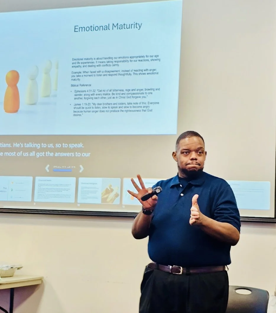 A man in a navy blue polo shirt giving a presentation on 'Emotional Maturity' in a classroom or conference room. He is making a hand gesture with his right hand and giving a thumbs-up with his left hand. A projection screen behind him displays text and images related to the presentation.