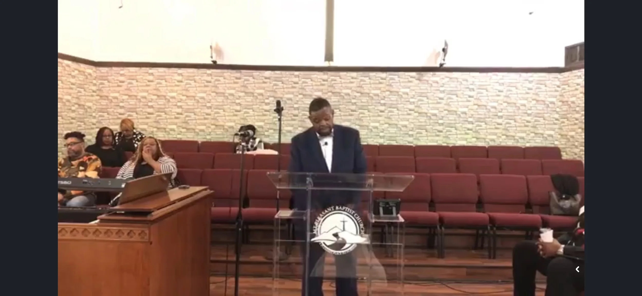 A man in a suit standing at a clear pulpit with a church emblem, speaking or praying at a church service. Several people are seated behind him on maroon chairs, some with their hands on their faces, and others with devices or drinks in their hands.