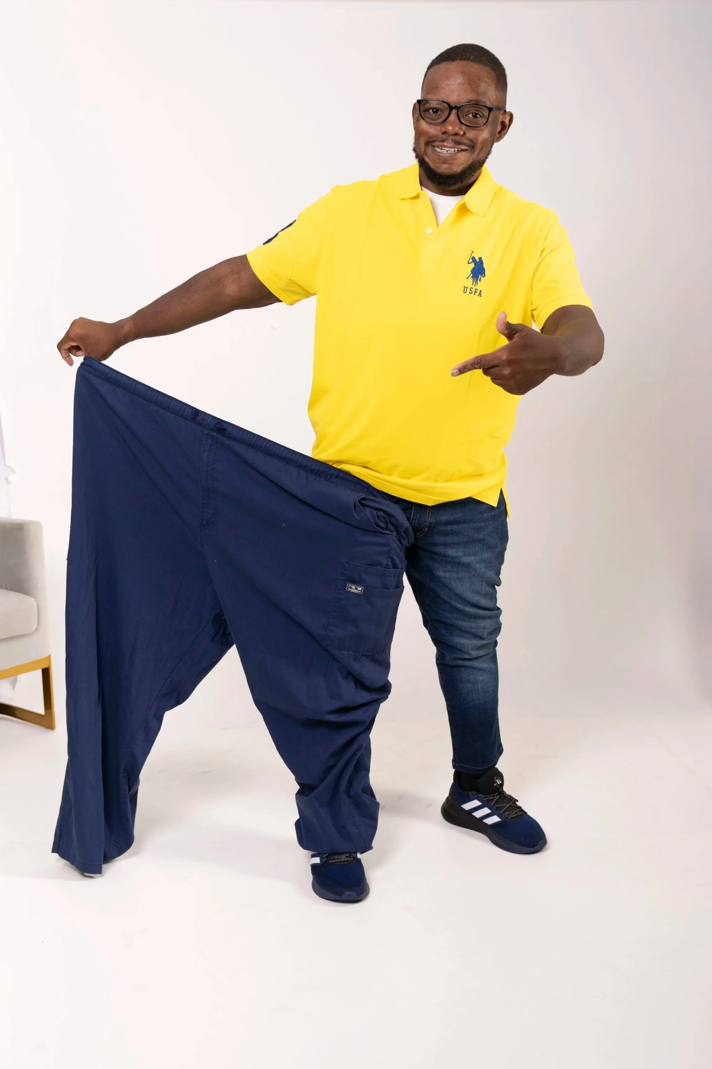 Man holding oversized pants and pointing at them, wearing a yellow polo shirt with a logo and glasses, smiling in studio