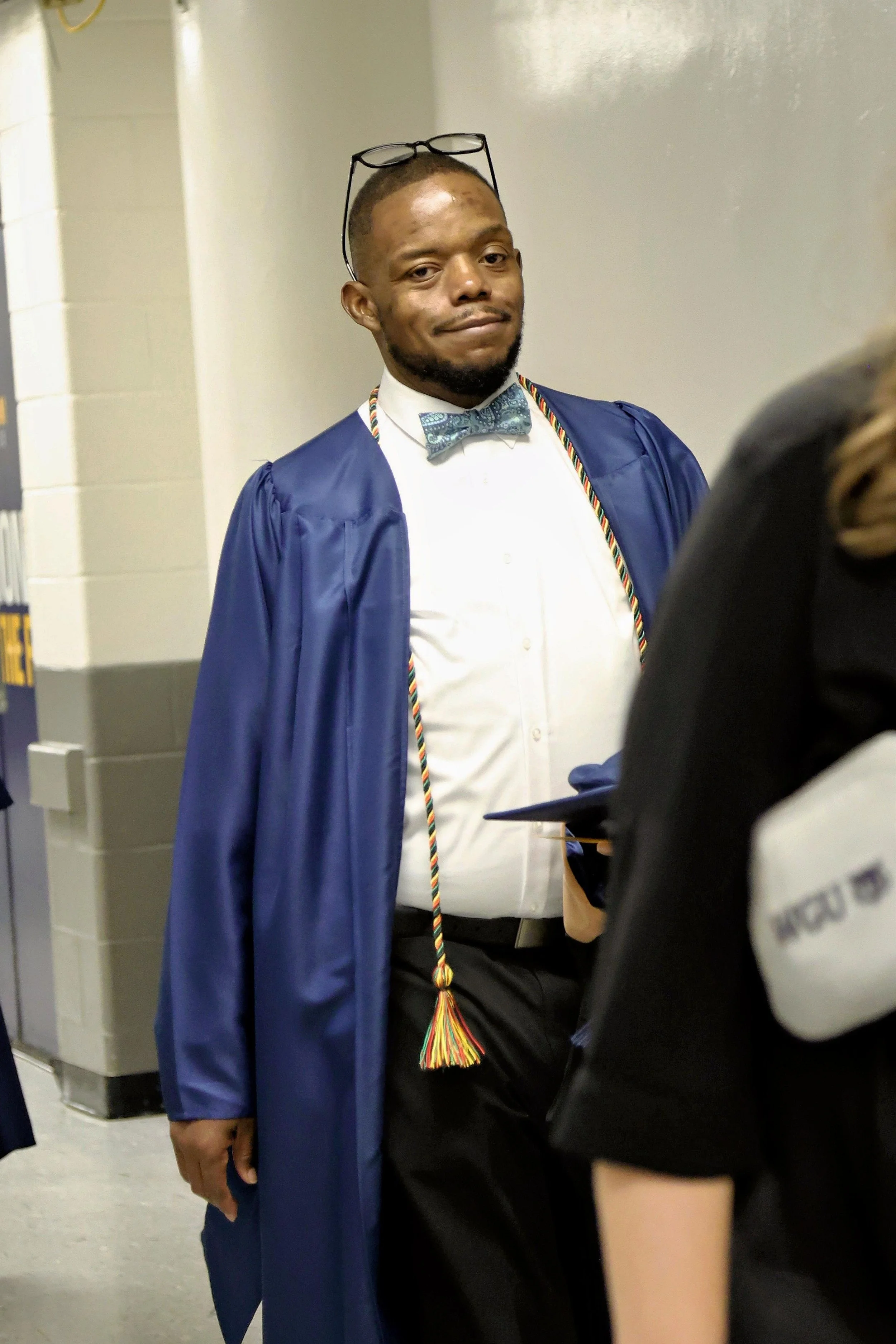 A young man in a blue graduation gown and cap with glasses resting on his head, wearing a white shirt and a bow tie, stands in a hallway.