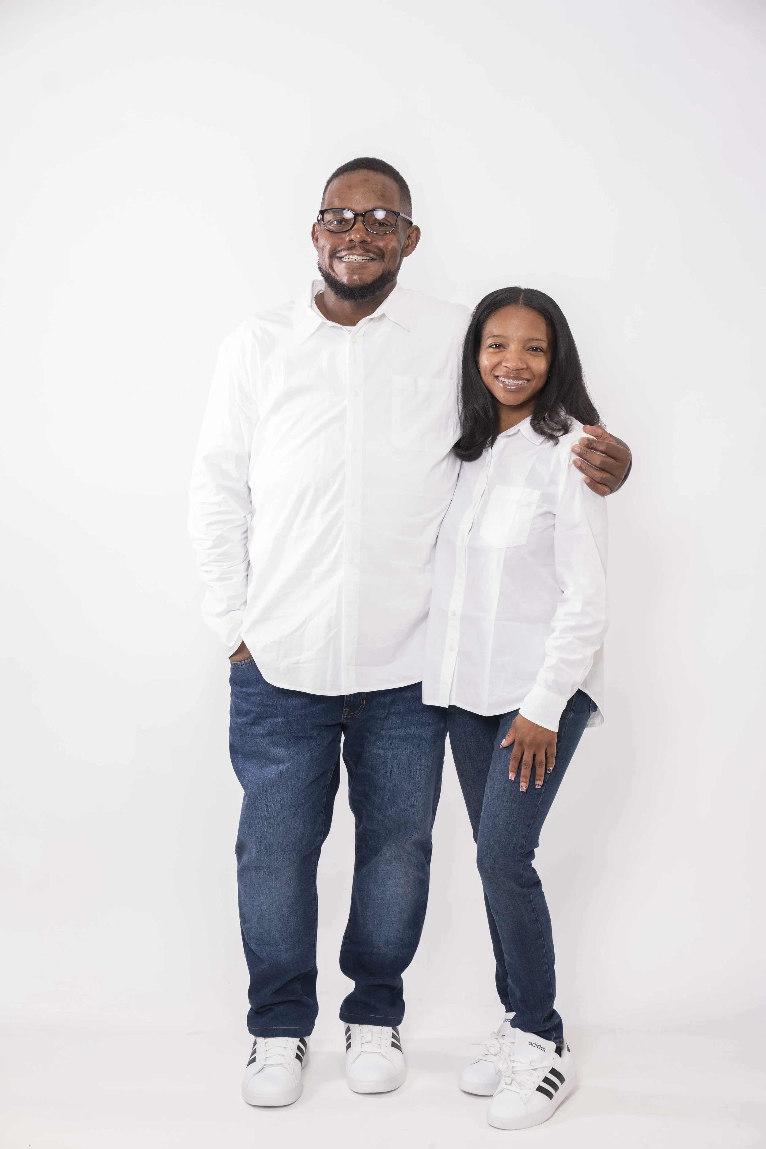 A smiling Black man and woman dressed in white shirts and jeans, standing close together with the man's arm around the woman's shoulder against a plain white background.