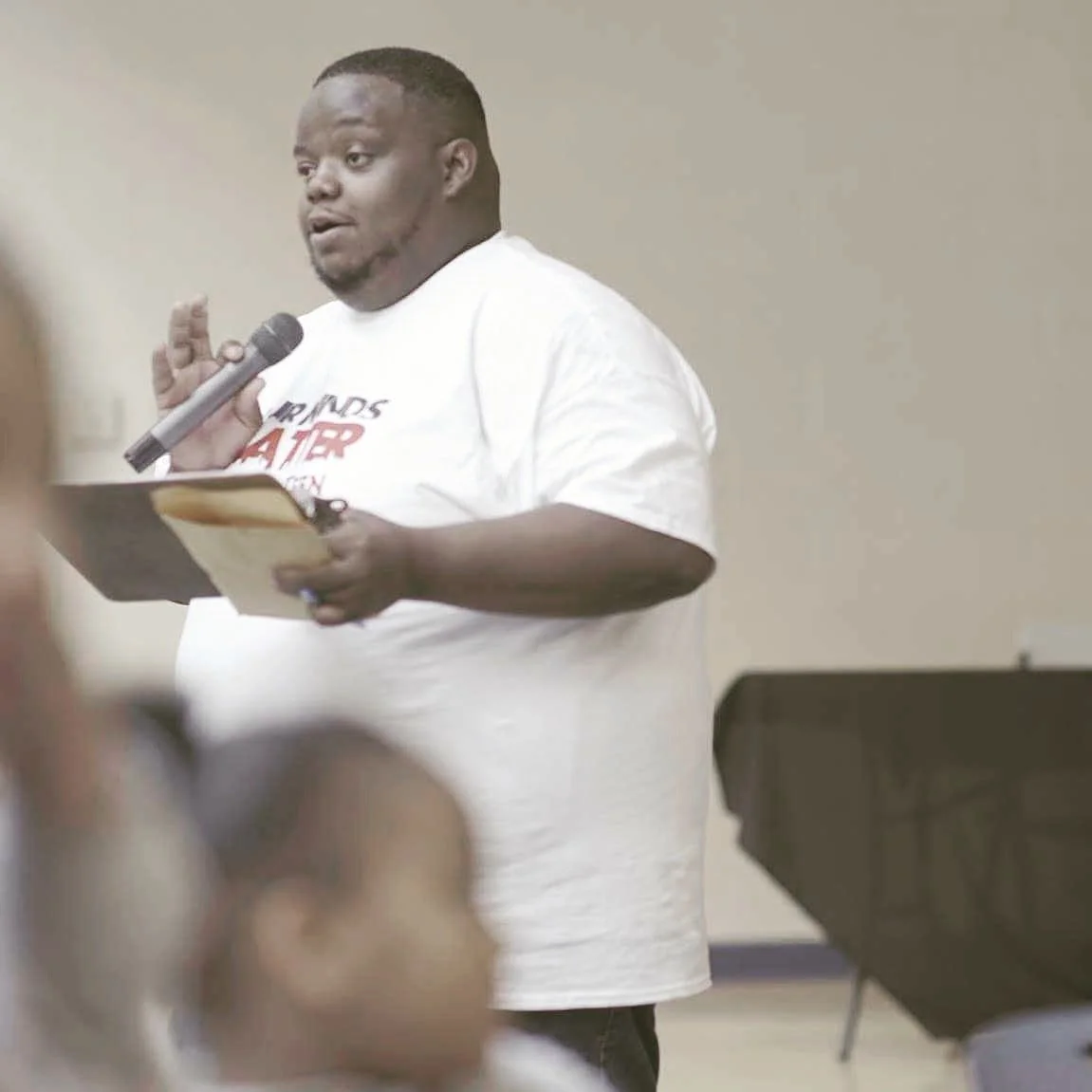 Man speaking into a microphone while holding a script or notes in a room with a podium and a beige wall.