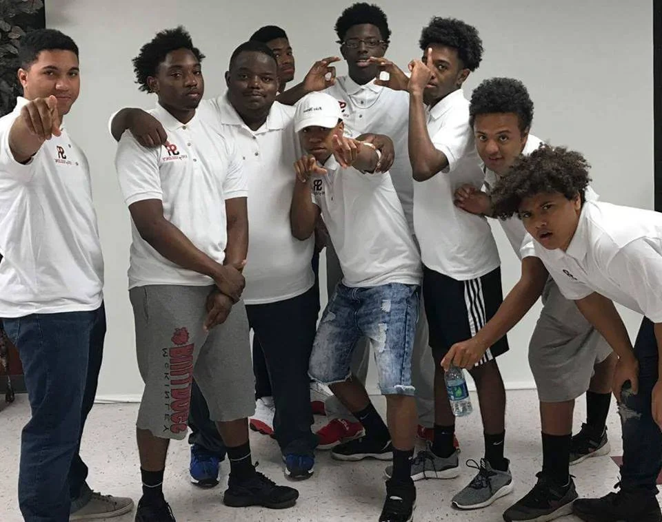 Group of nine young males posing together indoors, some making hand gestures, all wearing white polo shirts and athletic shorts or pants.