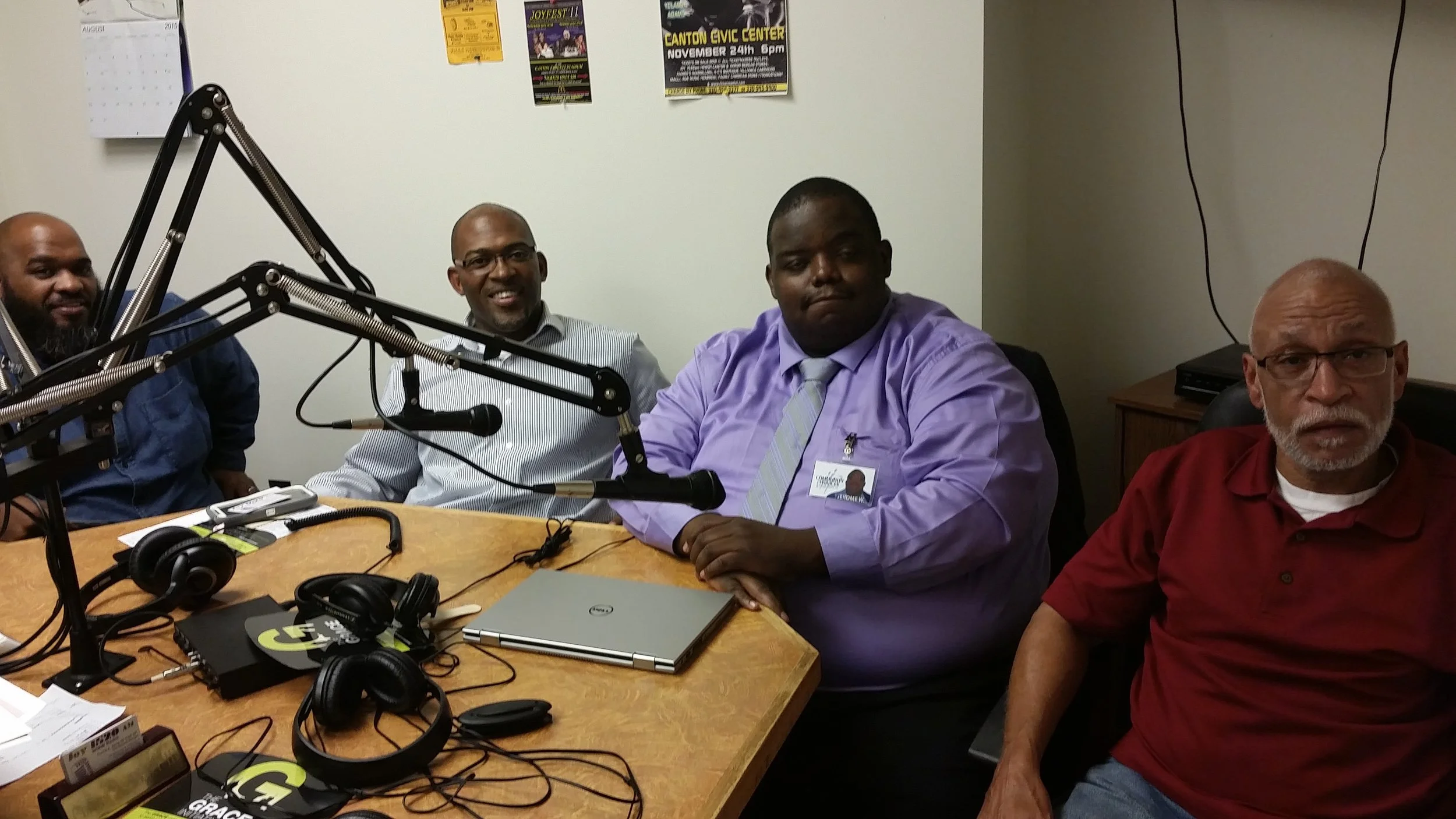 Four men sitting around a radio studio table with microphones and headphones, with posters on the wall behind them.