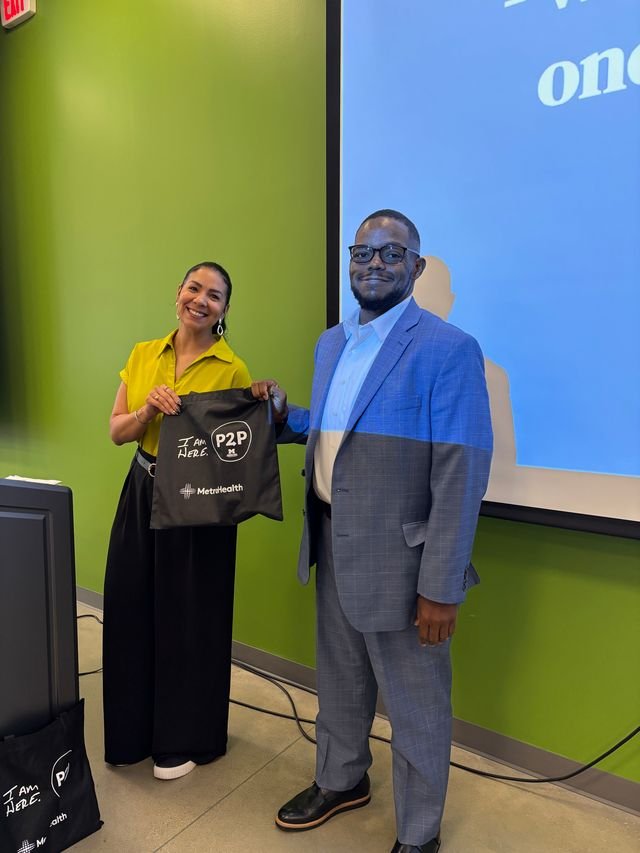 A woman in a yellow blouse and black pants and a man in a blue suit are standing indoors, smiling, with the woman holding a black tote bag that says "I AM HERE" and has P2P and MetroHealth logos. They are near a green wall and a large screen.