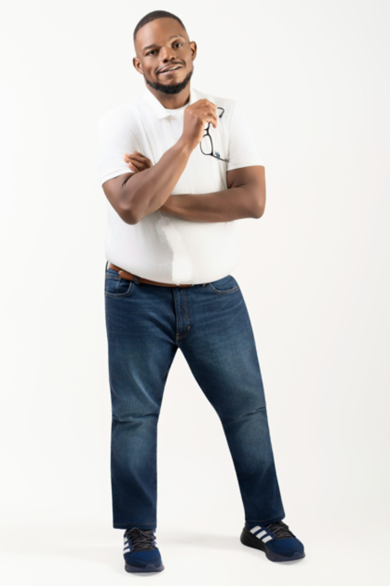 A confident man with a beard, wearing a white polo shirt, blue jeans, and sneakers, is holding glasses in his right hand and smiling at the camera.