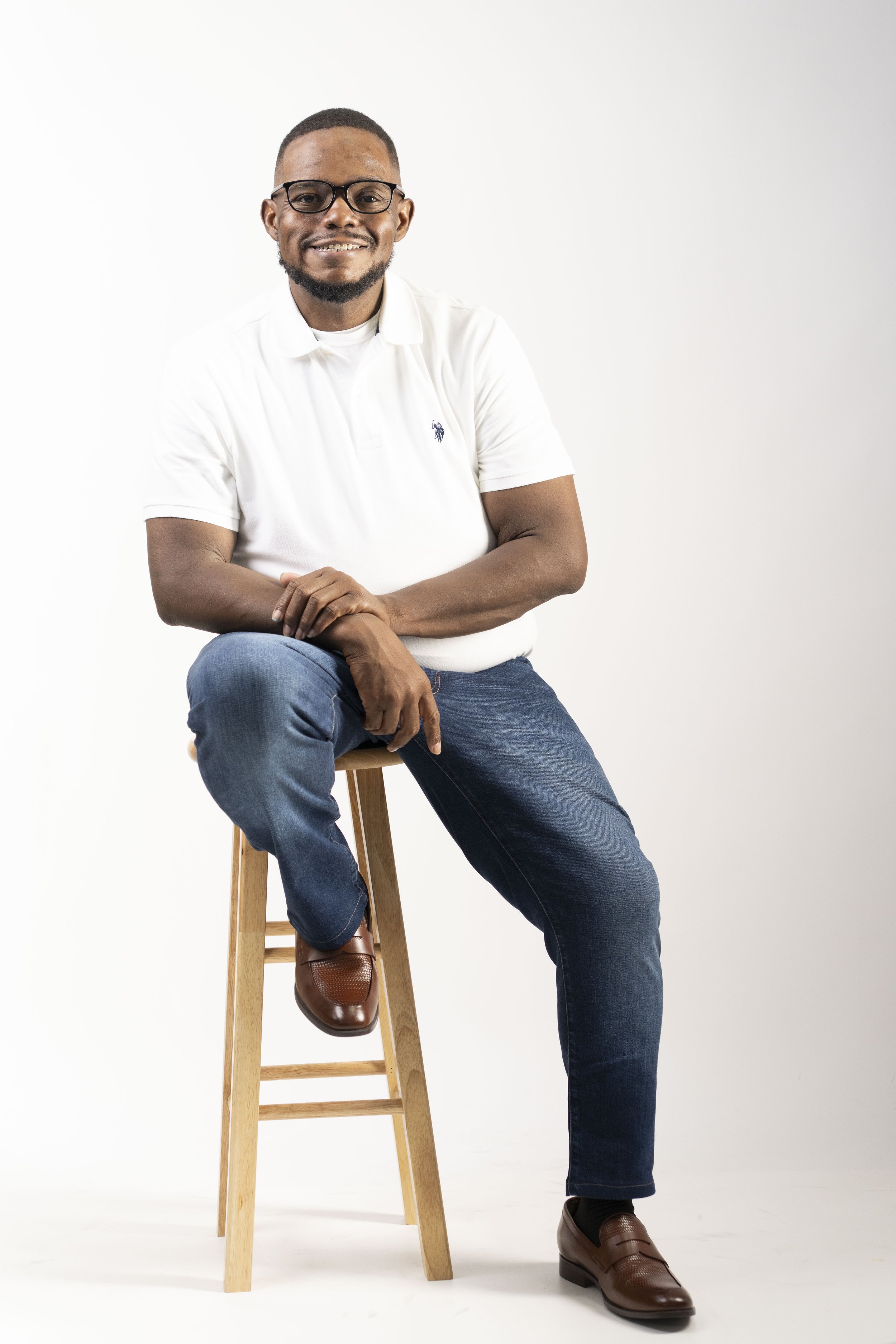 A smiling man with glasses and a beard sitting on a wooden stool against a plain white background. He is wearing a white polo shirt, blue jeans, and brown shoes, with arms crossed and one leg propped on the stool.