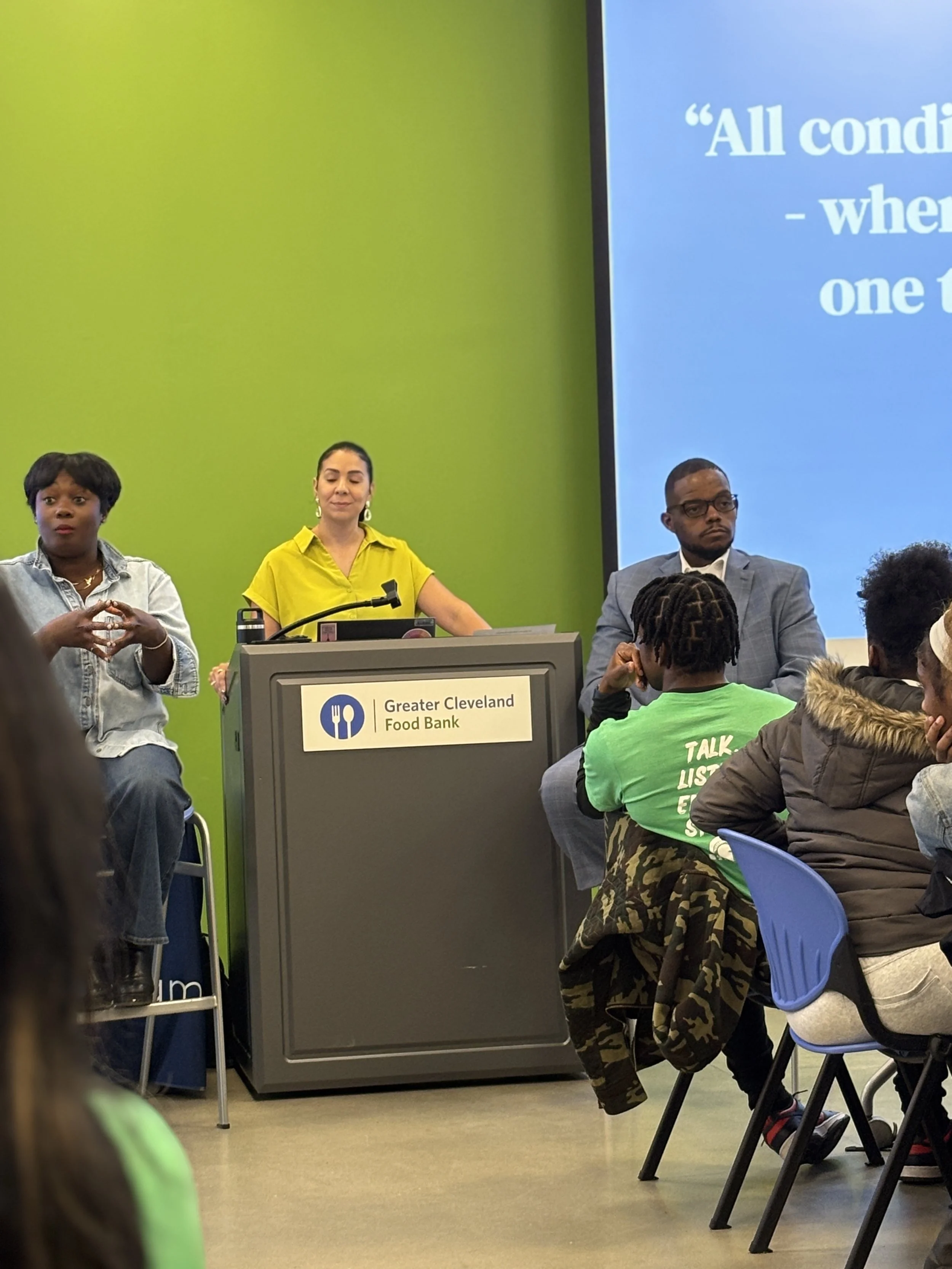 A panel discussion at Greater Cleveland Food Bank with three speakers seated at a table, including one woman in a bright yellow shirt, and an audience of people listening attentively.