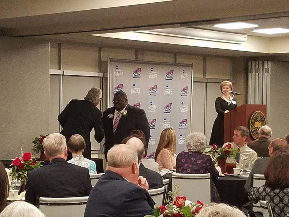 A woman standing at a podium speaking at a formal event, with people seated at tables adorned with flowers watching. Two people are shaking hands in front of a backdrop with logos.