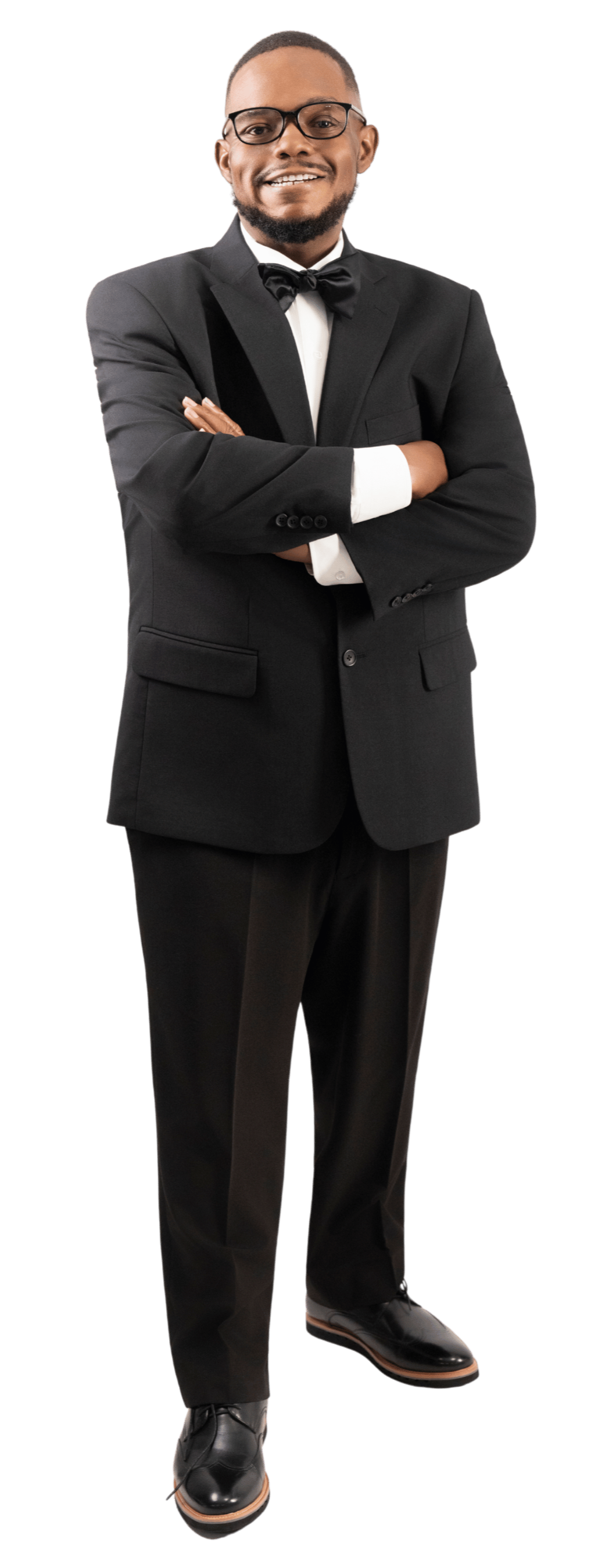 A man in a black tuxedo with a bow tie, wearing glasses, standing with his arms crossed and smiling.