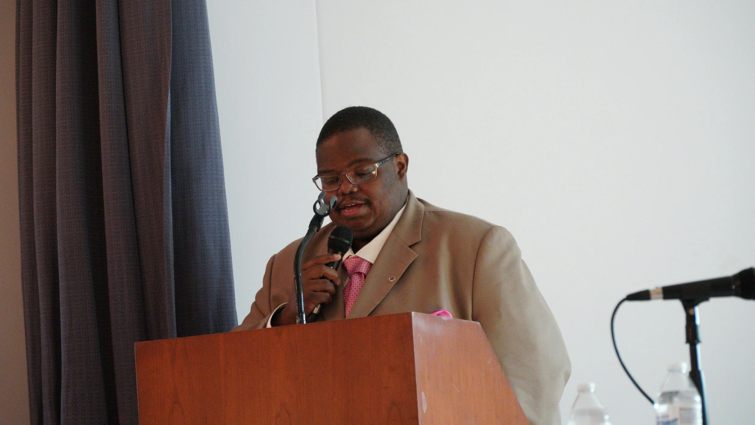 A man wearing a beige suit, pink tie, and glasses is speaking into a microphone at a wooden podium. There are three water bottles on a table nearby and a microphone on a stand to the right. The background consists of a white wall and dark curtains.