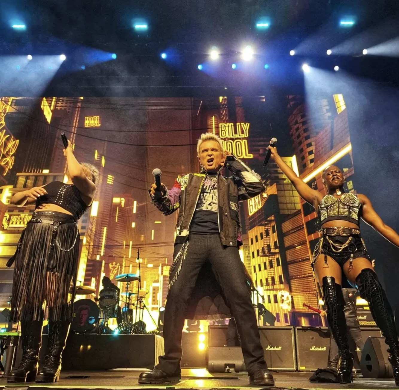 Musical performance on stage with three singers, one male in the center and two women on each side, against a cityscape backdrop with yellow lighting and the words 'Billy Kool' displayed.