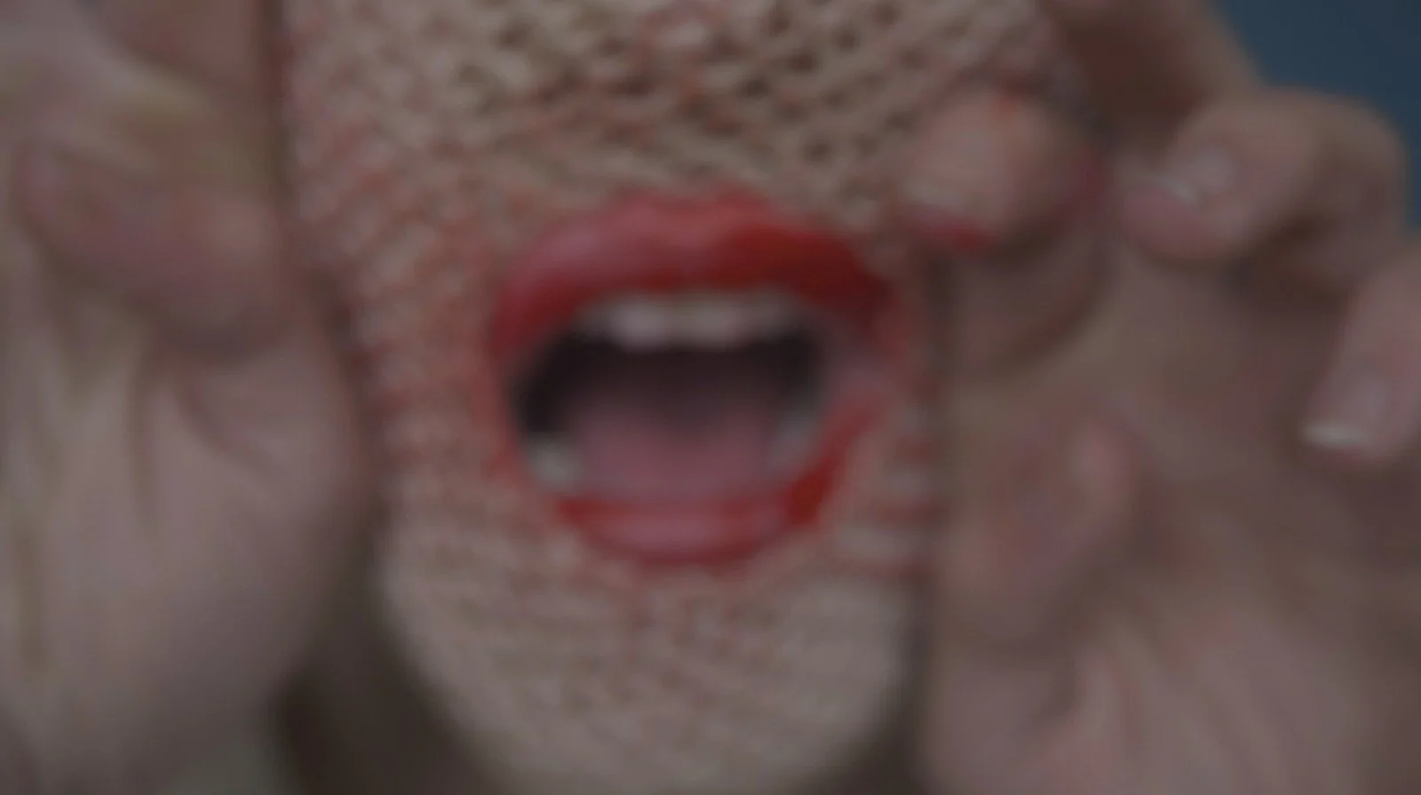 Close-up of a person's face with red lipstick, showing an open mouth, hands near the cheeks, and a textured background.