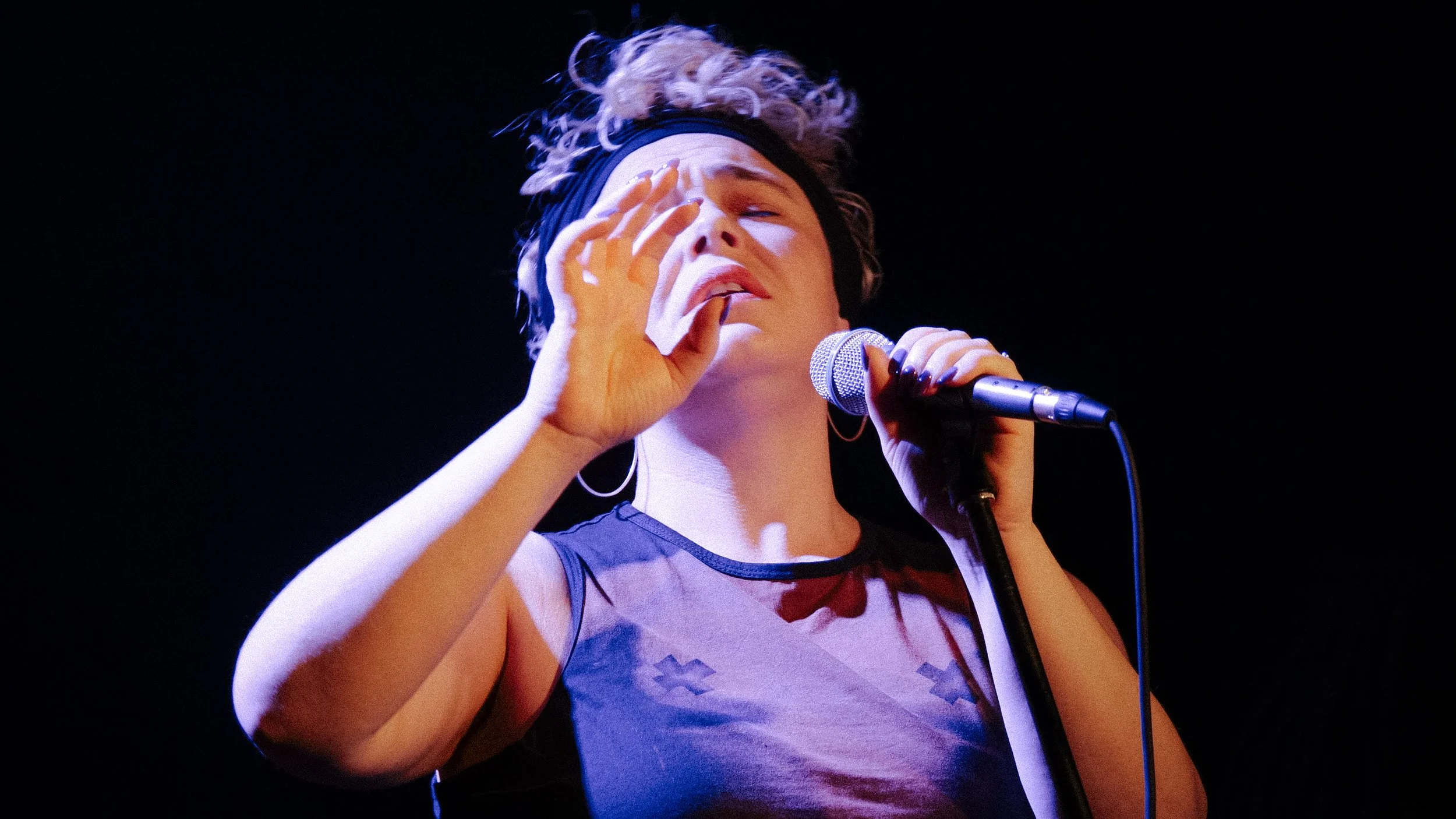 A woman with curly hair, wearing a headband, singing into a microphone on stage with a black background.