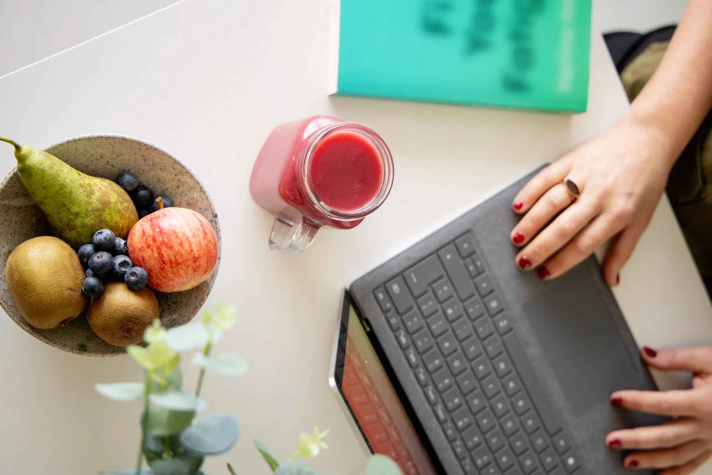 A person using a laptop on a white table with a bowl of assorted fruits, a glass of pink smoothie, and some books.