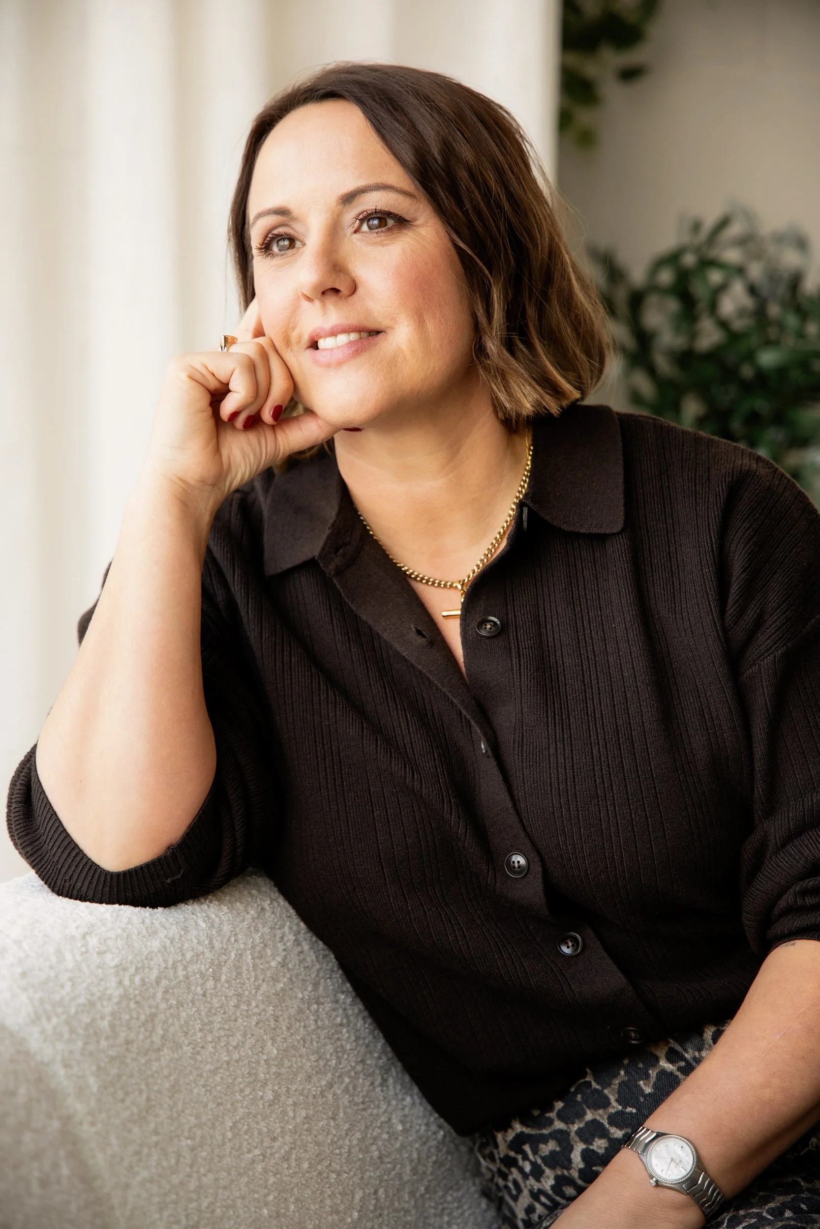 A woman with short brown hair and fair skin, wearing a black button-down shirt, a gold necklace, and a silver watch, is resting her chin on her hand and smiling softly in a well-lit indoor setting.