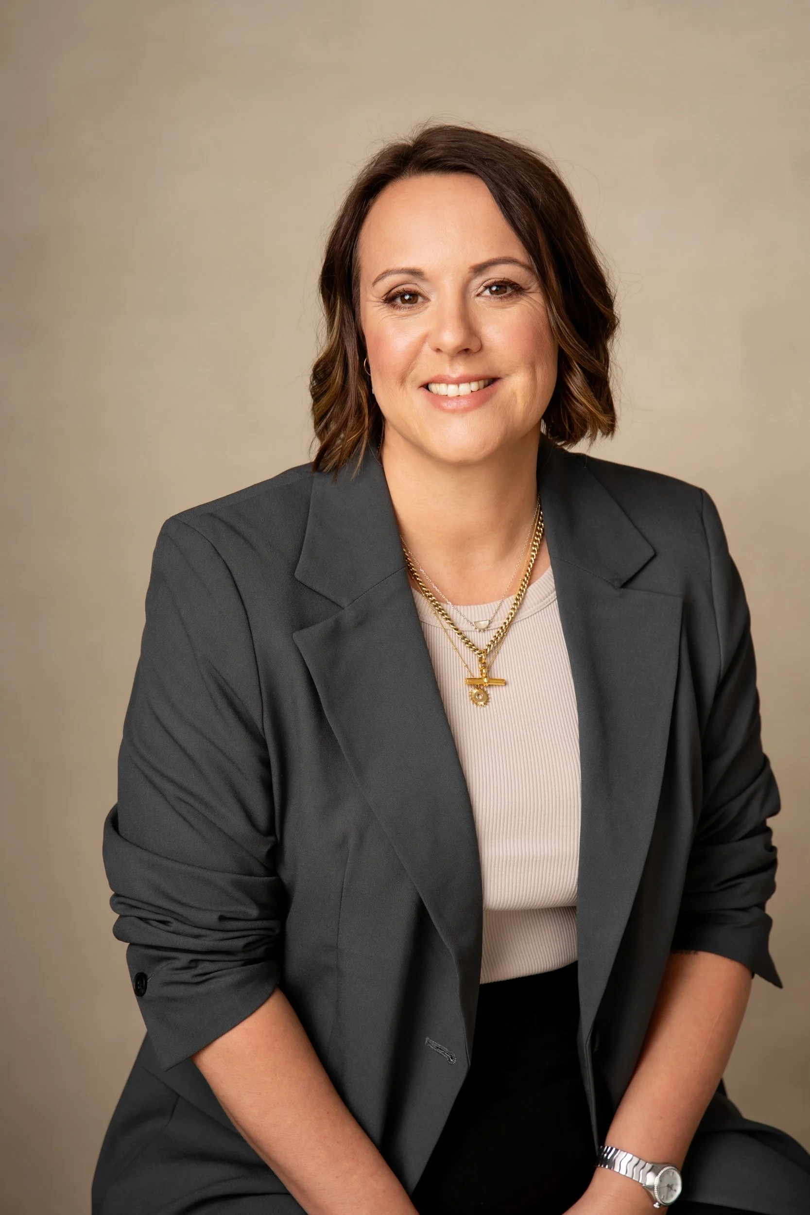 A woman with short brown hair, wearing a dark gray blazer and a beige top, smiling at the camera against a neutral background.