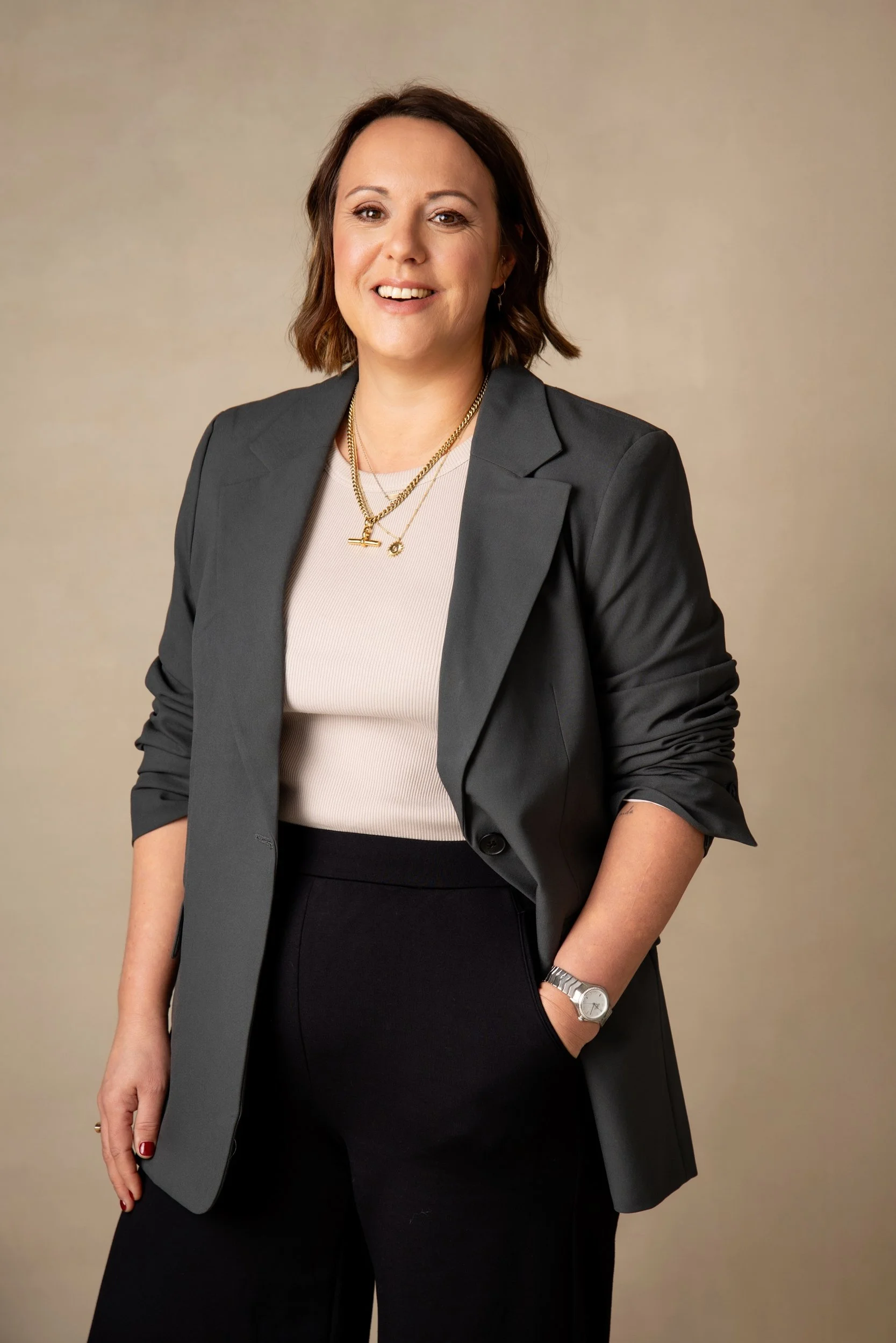 A woman with shoulder-length brown hair smiling, wearing a dark gray blazer over a white top, black pants, a silver watch, and layered gold necklaces, standing in front of a beige background.