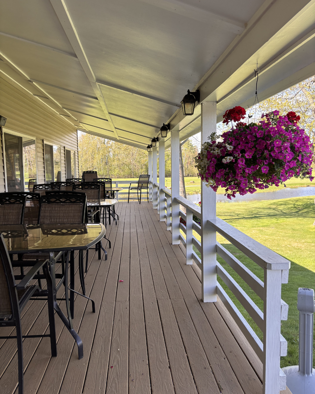 A spacious outdoor porch with multiple tables and chairs arranged for dining, a hanging basket of colorful pink and white petunias, white wooden railing, and a scenic view of a pond and green lawn in the background.