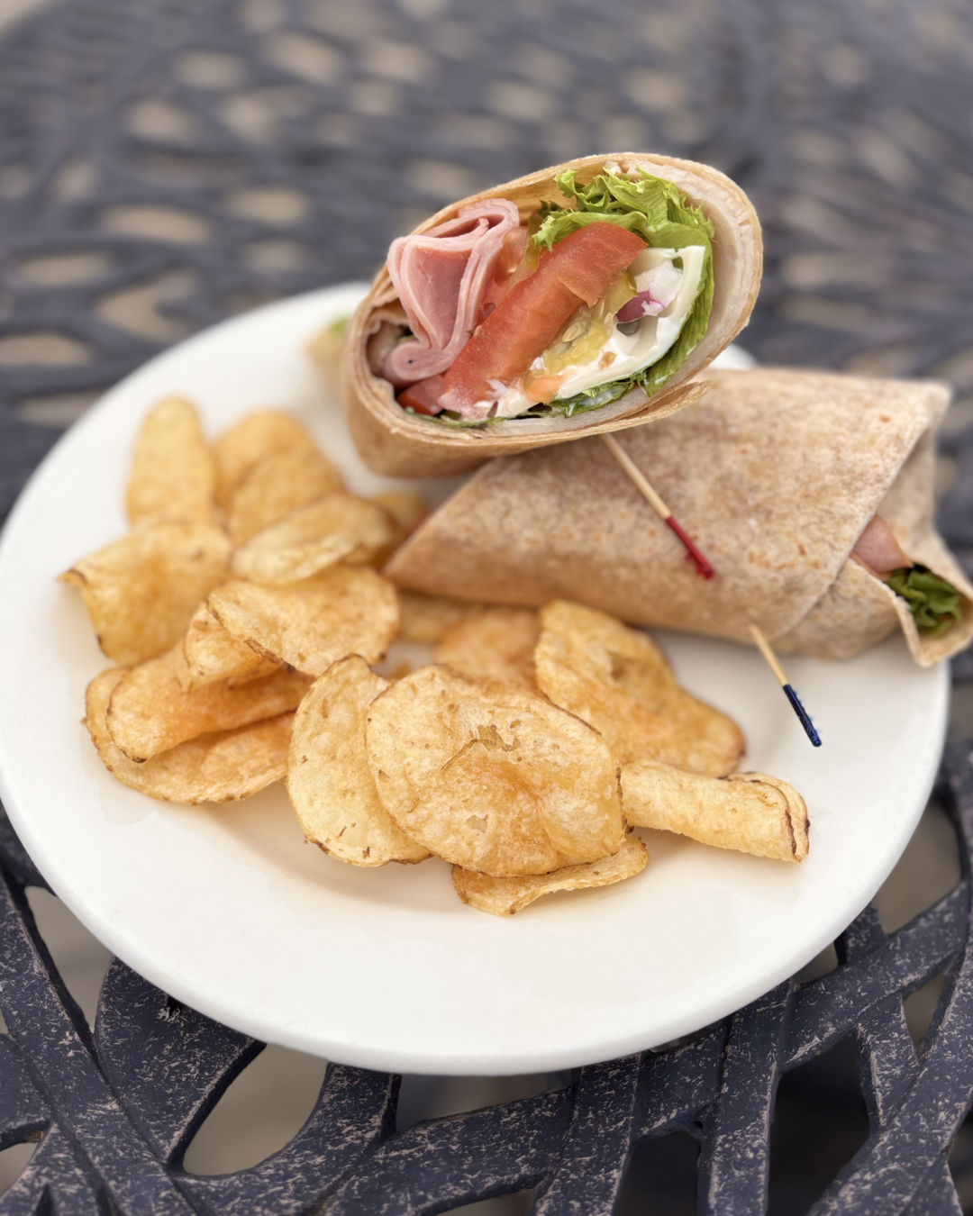 A plate with potato chips, a wrapped sandwich, and a wrap snack, with a sandwich cut in half showing ham, lettuce, tomato, and other vegetables.