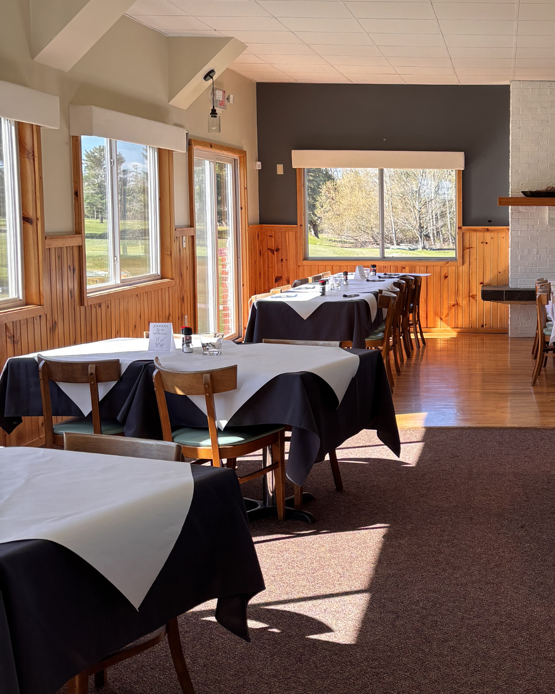 A sunlit dining room with multiple tables covered in black and white tablecloths. Wooden chairs surround the tables, and large windows offer a view of a sunny outdoor landscape with trees and grass. The room has wooden paneling on the lower half of t