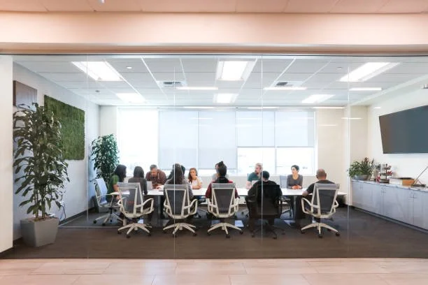Five people sitting at a conference table in a modern office, engaged in a discussion, viewed through a glass wall.