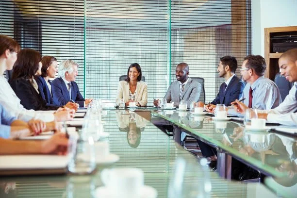 Business meeting in a conference room with a presenter and attendees seated around a table with laptops, gift bags, and notebooks.