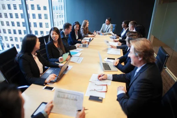 Business meeting in a conference room with a presenter and attendees seated around a table with laptops, gift bags, and notebooks.