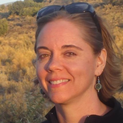 A woman with short brown hair, sunglasses on her head, and earrings, standing outdoors in a natural landscape with hills and trees, smiling at the camera during sunset.