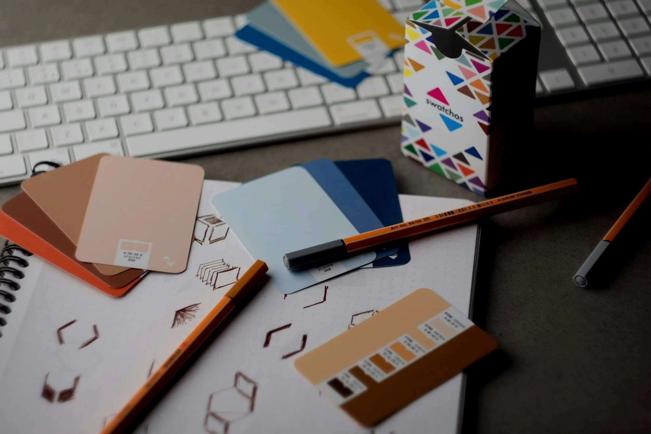 Color swatches, a sketchbook with geometric designs, markers, and a keyboard on a desk.