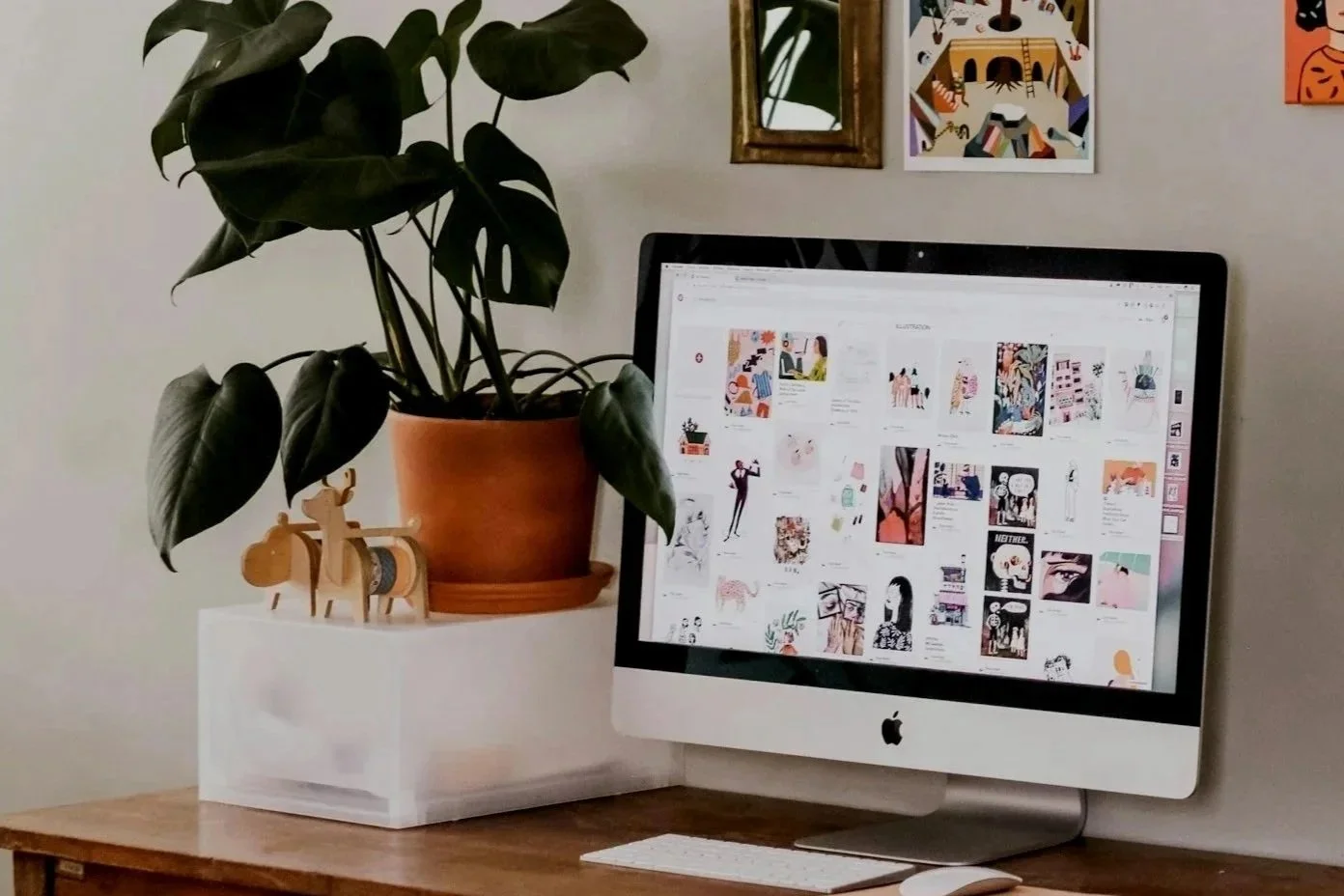 A computer monitor displaying a Pinterest page with various illustrations and artworks. To the left of the monitor, there is a potted plant with large green leaves in an orange terracotta pot on top of a white container. Next to the plant, there are two wooden figurines. On the wall above the computer, there are three framed pictures, including a mirror and colorful artwork.