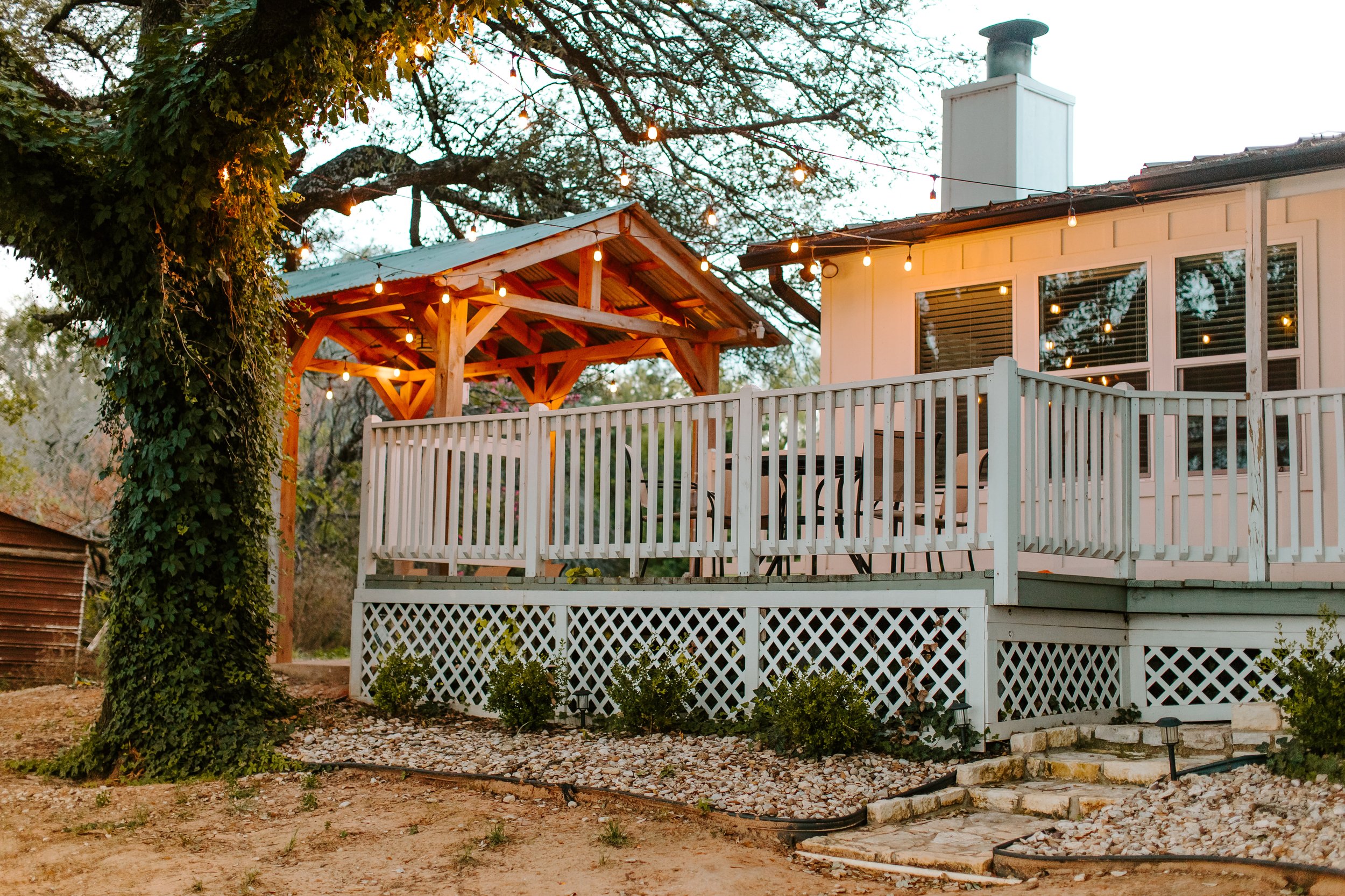 A backyard deck with string lights hanging above, surrounded by trees and plants, with a white railing and a wooden pergola with string lights.