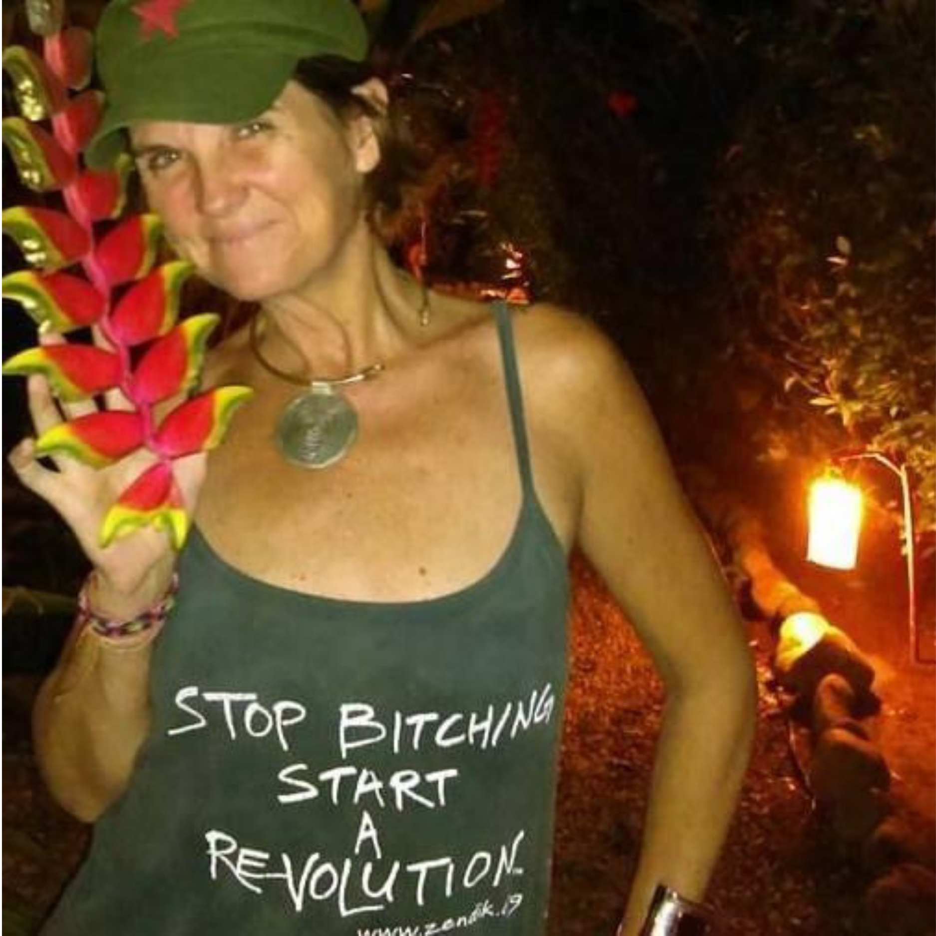 Person holding a colorful flower lei at night with a sign that reads 'Stop Bitching, Start Rebellion'.