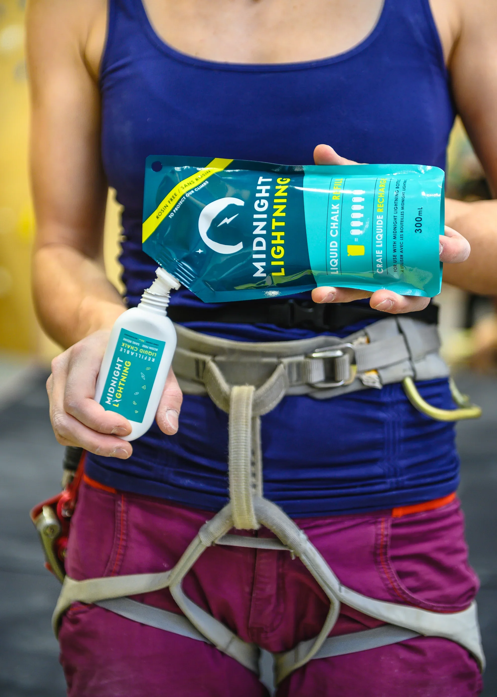 A person wearing a blue tank top and purple climbing pants holding a bottle and a pouch of Midnight Lightning liquid chalk for rock climbing.