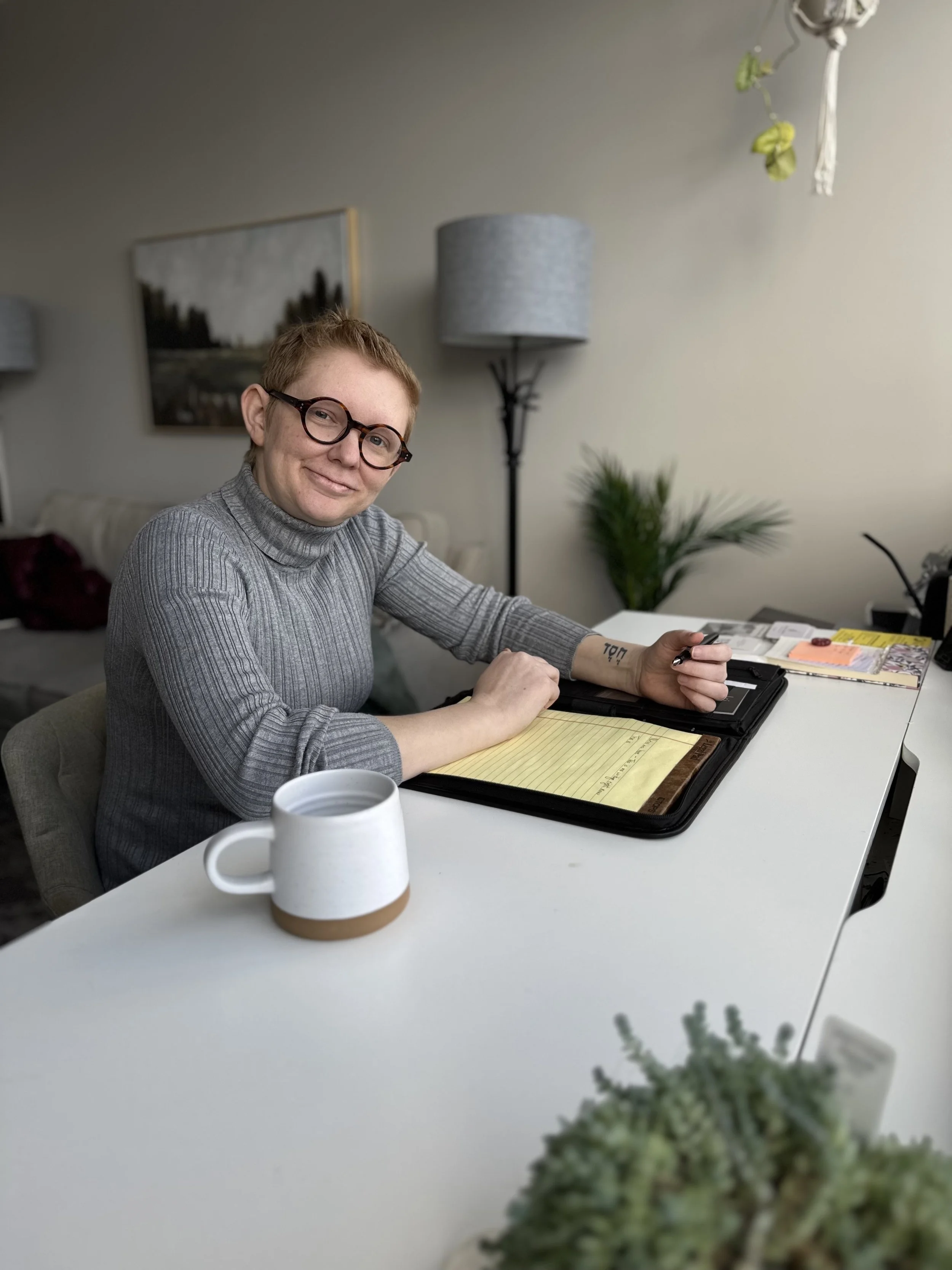 Portrait of a woman with short hair and glasses smiling while sitting at a white table with documents, a mug, and a pen in a cozy room with a plant, a floor lamp, and artwork on the wall.