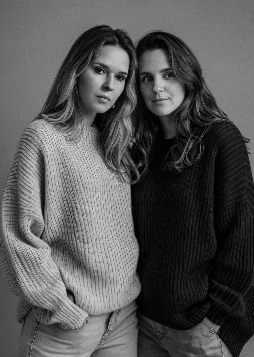 Duas mulheres jovens com cabelos longos, uma de cabelo loiro e outra de cabelo castanho, posando juntas em um fundo cinza, ambas usando suéteres de manga comprida, uma de cor clara e outra escura.