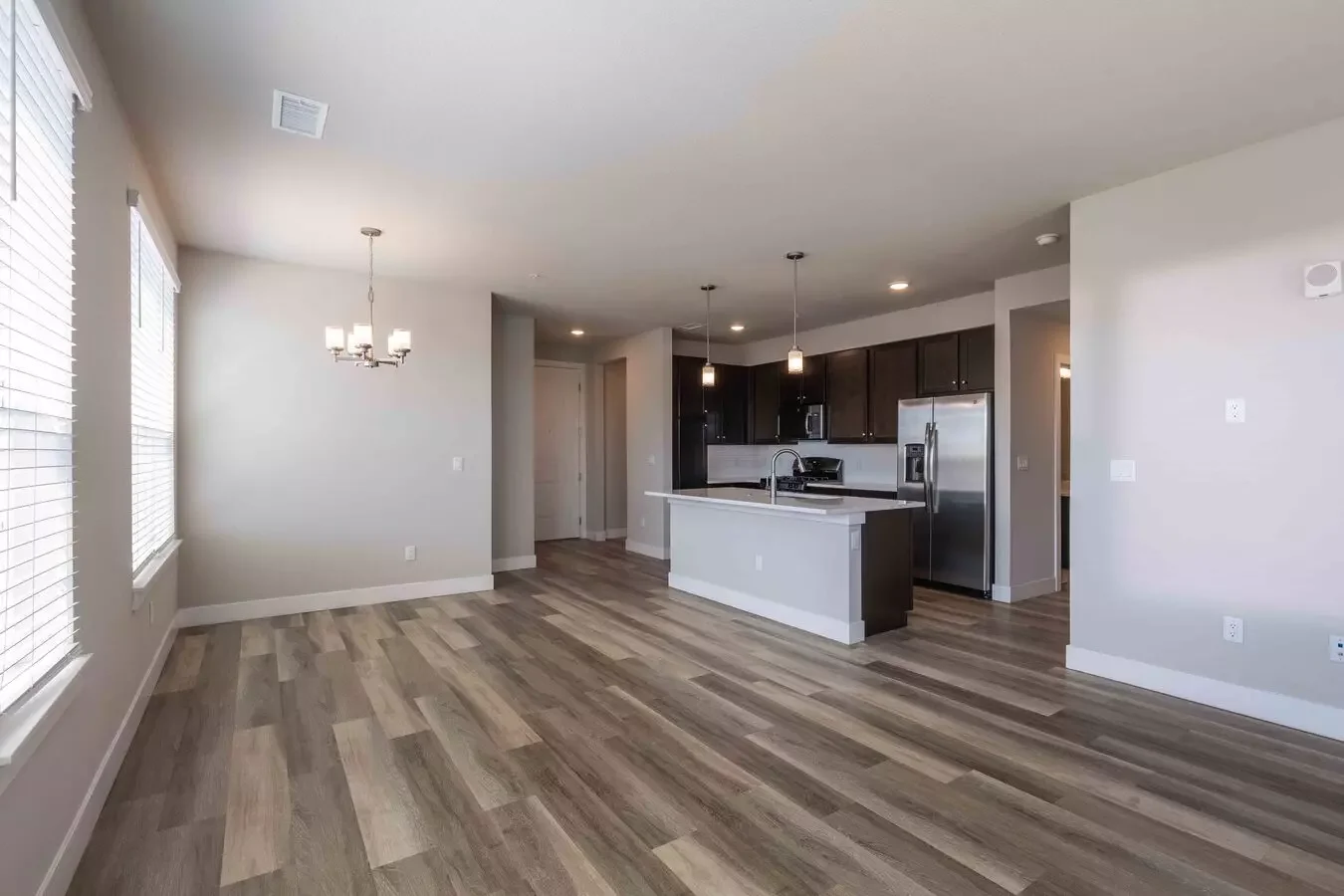 Empty living and kitchen area in a modern apartment with light wood flooring, white walls, and dark kitchen cabinets. The space features large windows with blinds, a chandelier, pendant lights, and stainless steel appliances.