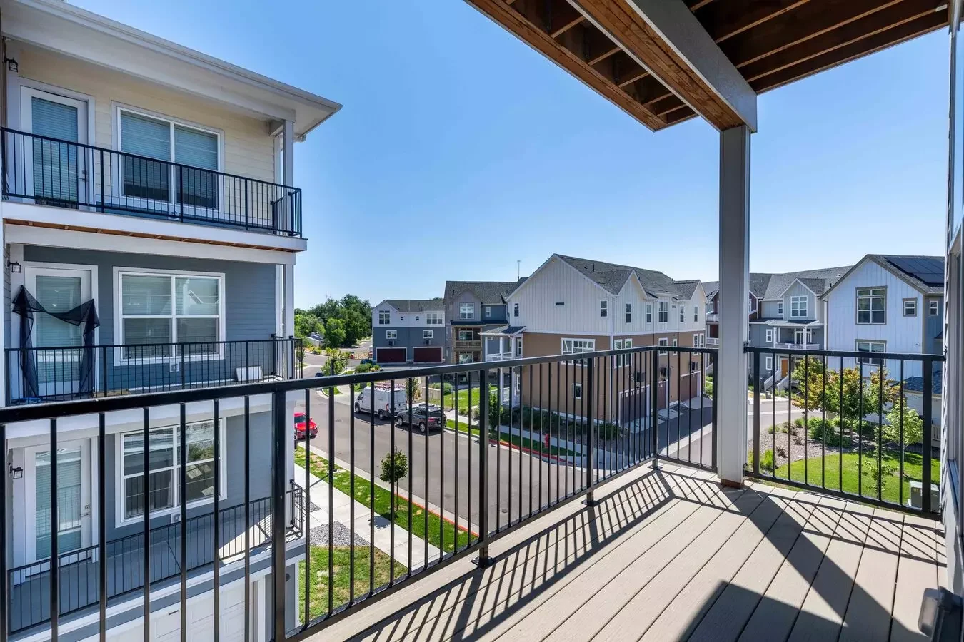 View from a balcony overlooking a residential neighborhood with modern multi-story houses, parked cars, and a bright blue sky.