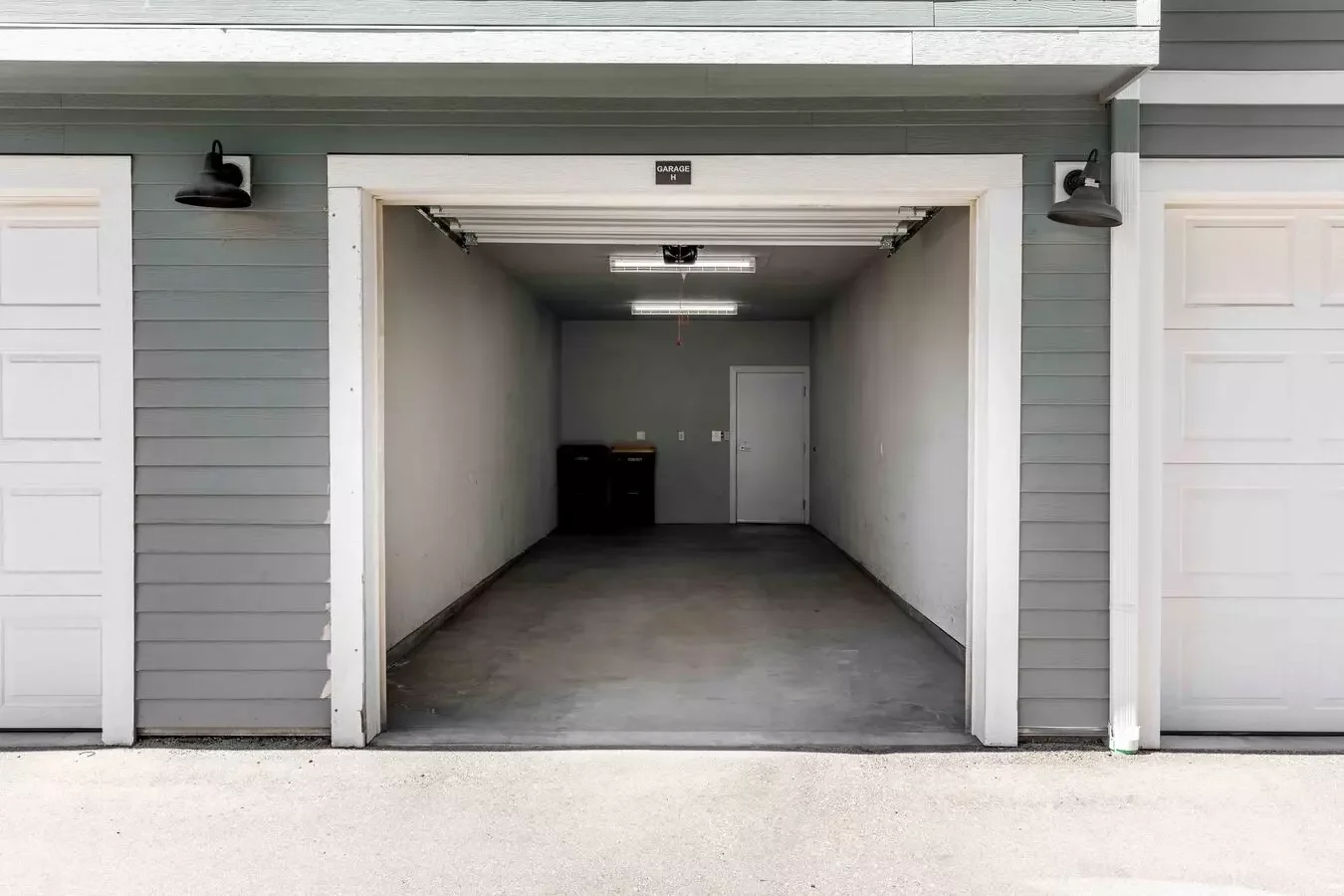 Open empty garage with gray interior walls and a door at the back, two black exterior lights above the garage opening, and a gray exterior siding with a small sign that reads Garage.