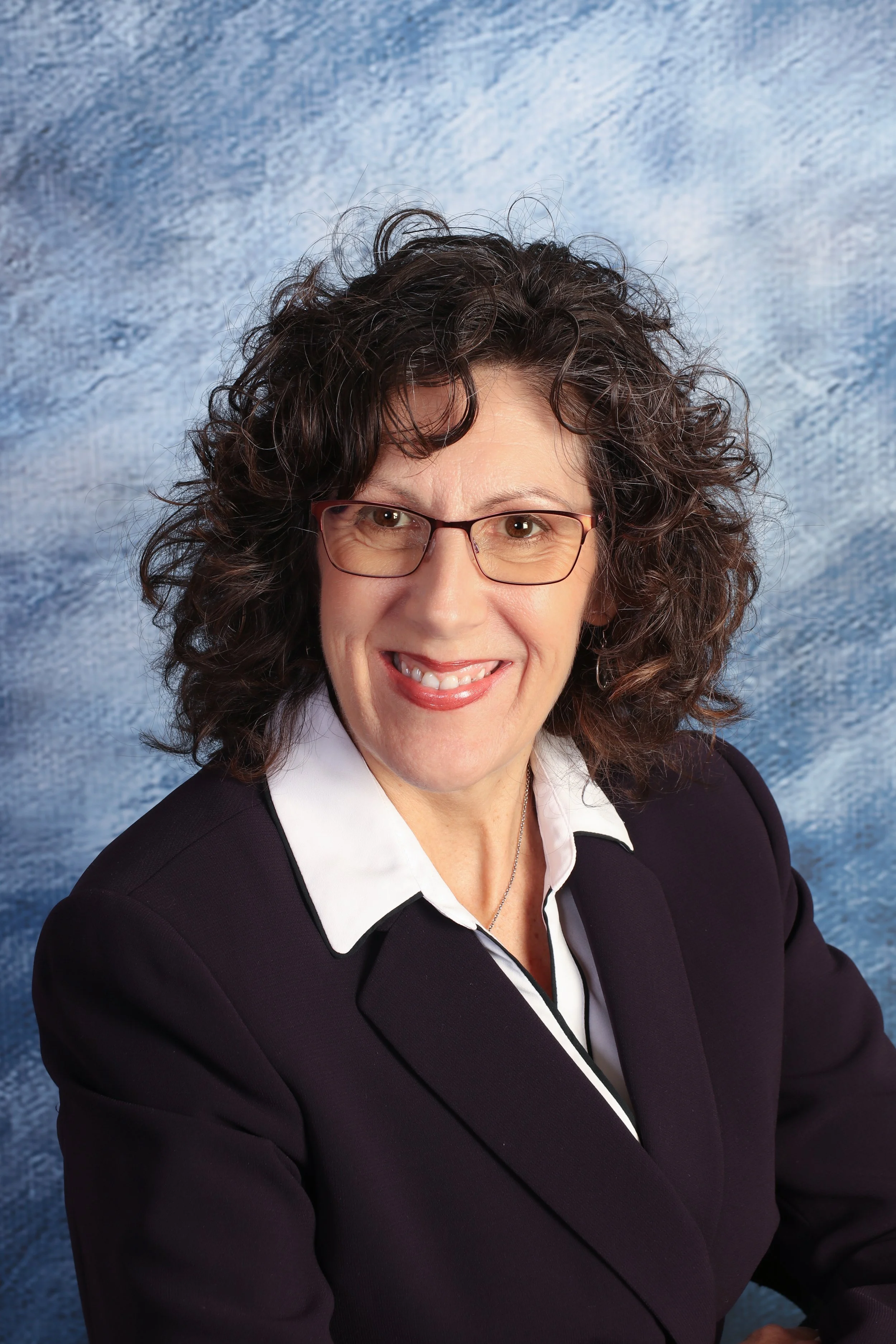 Portrait of Amy Irvine with curly dark hair and glasses, wearing a black blazer and white shirt, smiling against a blue textured background.