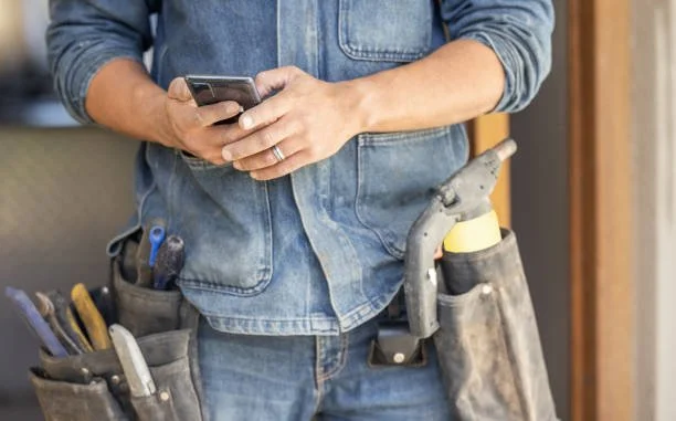 Person wearing denim overalls with a tool belt, holding a smartphone.