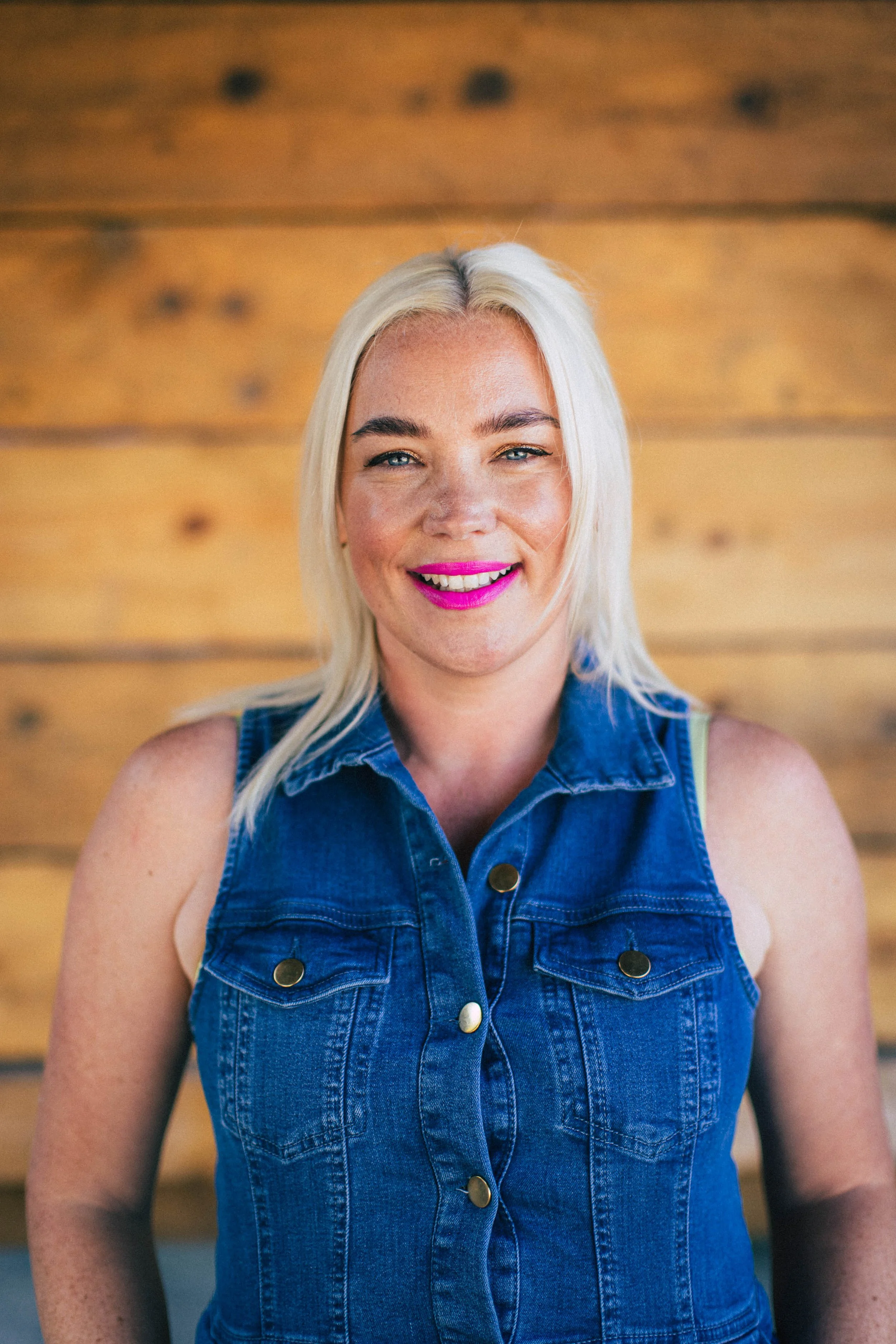 A woman with platinum blonde hair wearing a sleeveless denim vest, smiling in front of a wooden wall background.