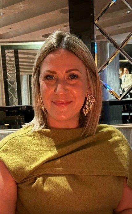 A smiling woman with long gray hair wearing a blue T-shirt in front of a wooden background.